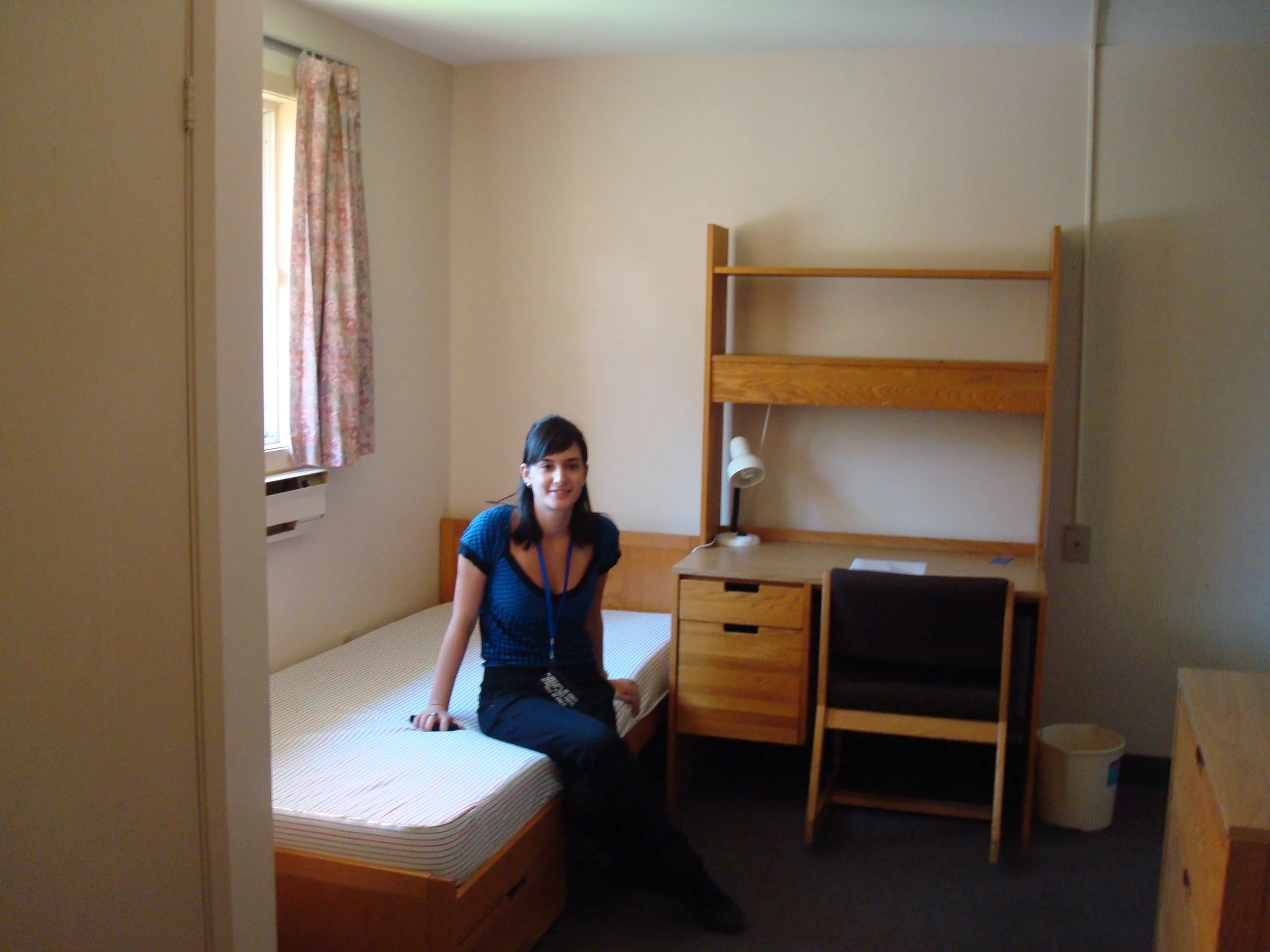 A young woman sitting on a bed in a dorm room with wooden furniture, a desk lamp, and a window with floral curtains.