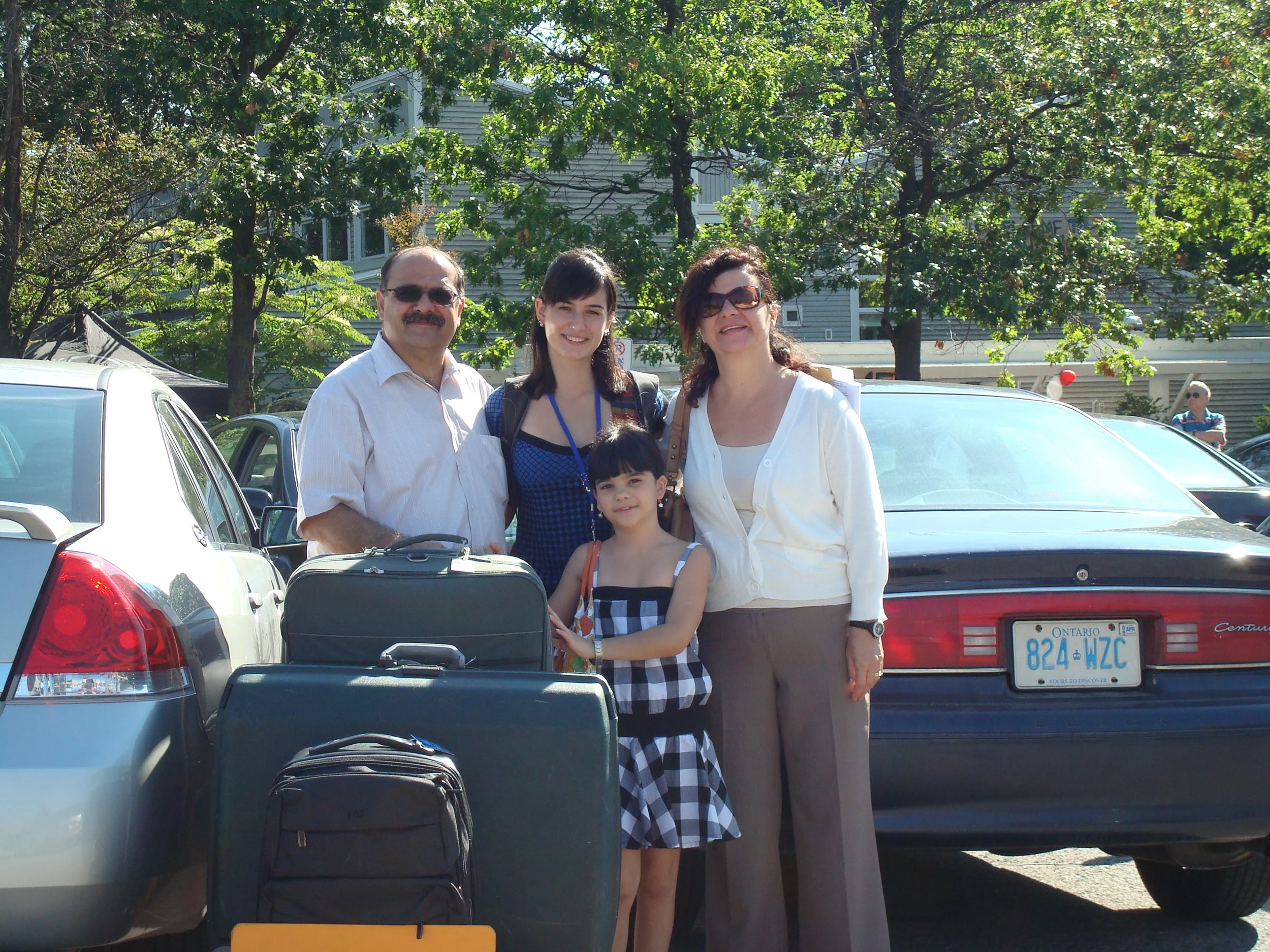 Four people standing together in a parking lot, with cars and trees in the background. It appears to be a family or group preparing for travel, with luggage and backpacks in front.