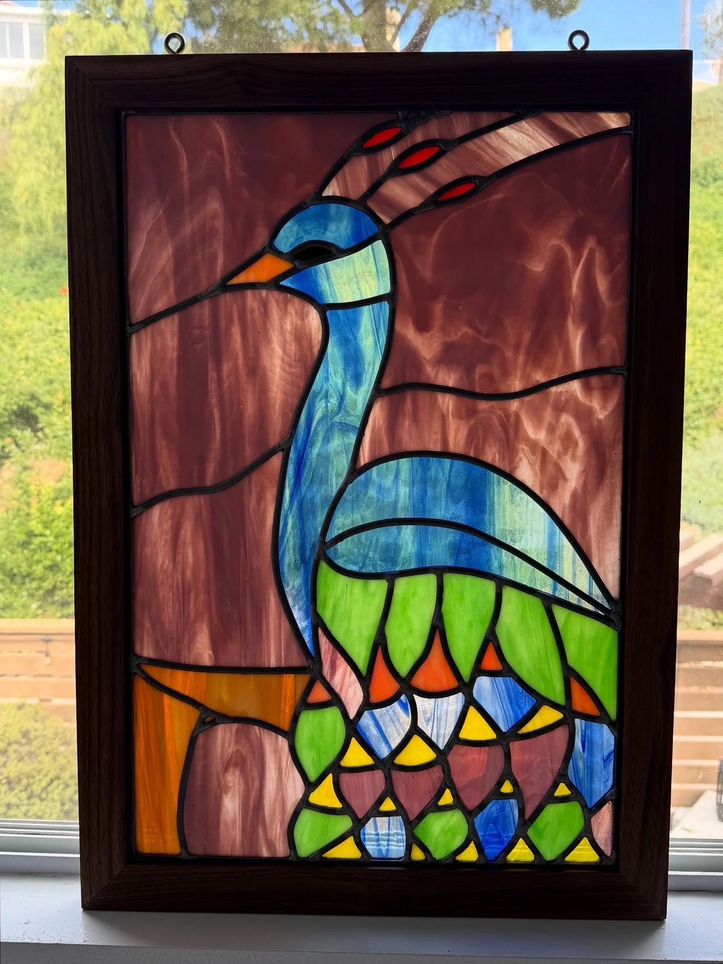 Colorful stained glass window depicting a peacock with blue, green, orange, yellow, and red glass segments, set in a wooden frame.