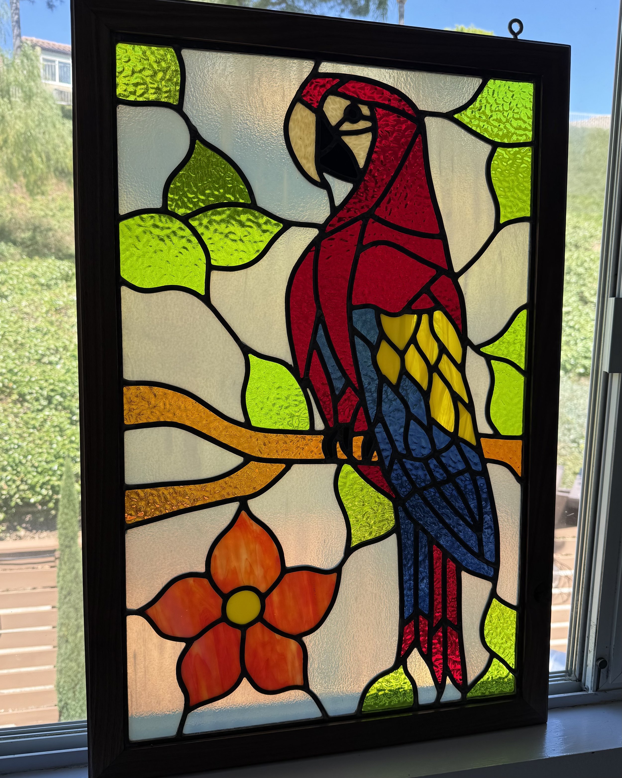 Colorful stained glass window featuring a bird with a red body, yellow wings, and a long blue tail, surrounded by green, yellow, white, and orange decorative glass pieces.