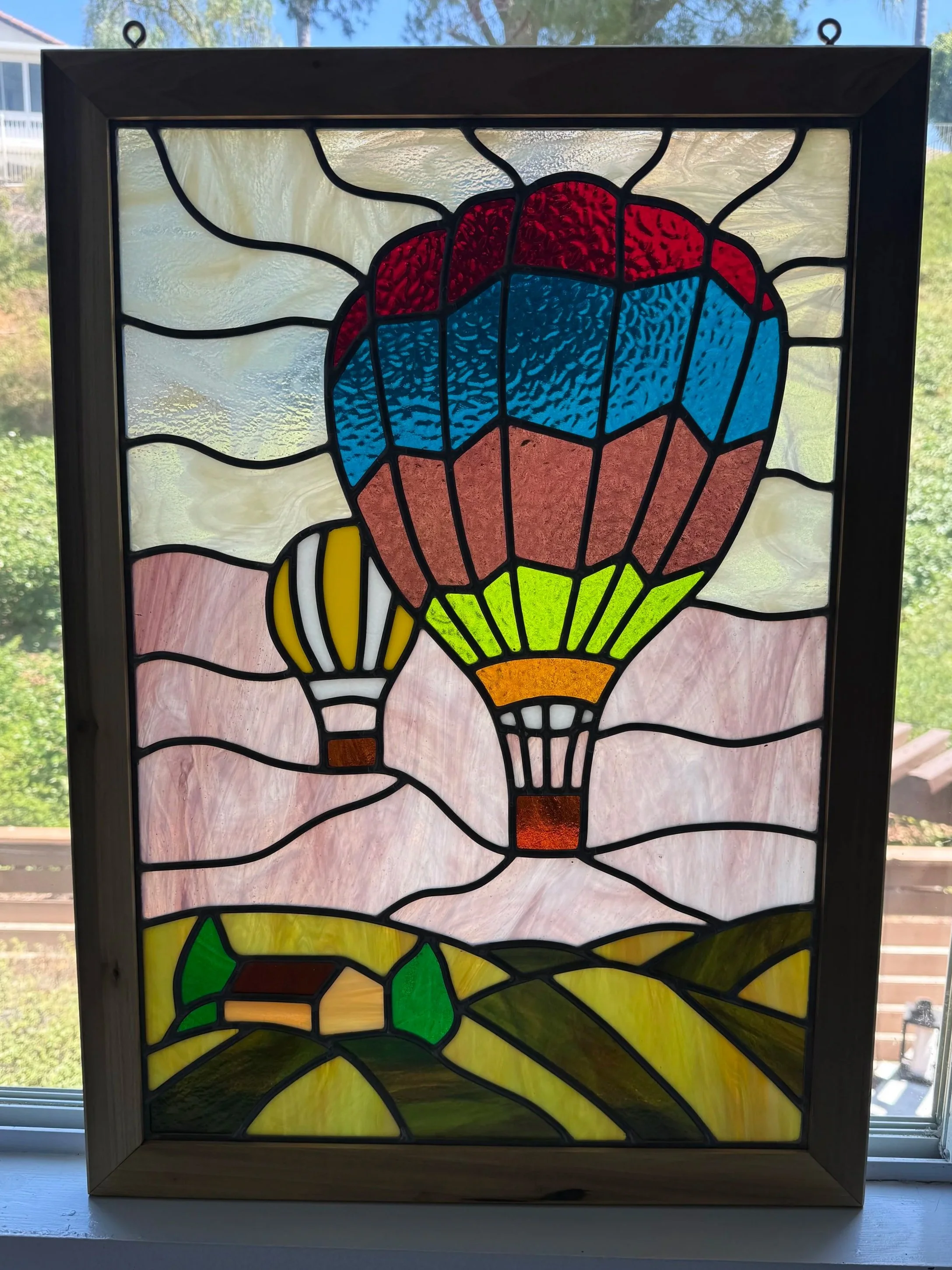 Stained glass window depicting a hot air balloon with a red, blue, and yellow design set against a cloudy sky and landscape background.
