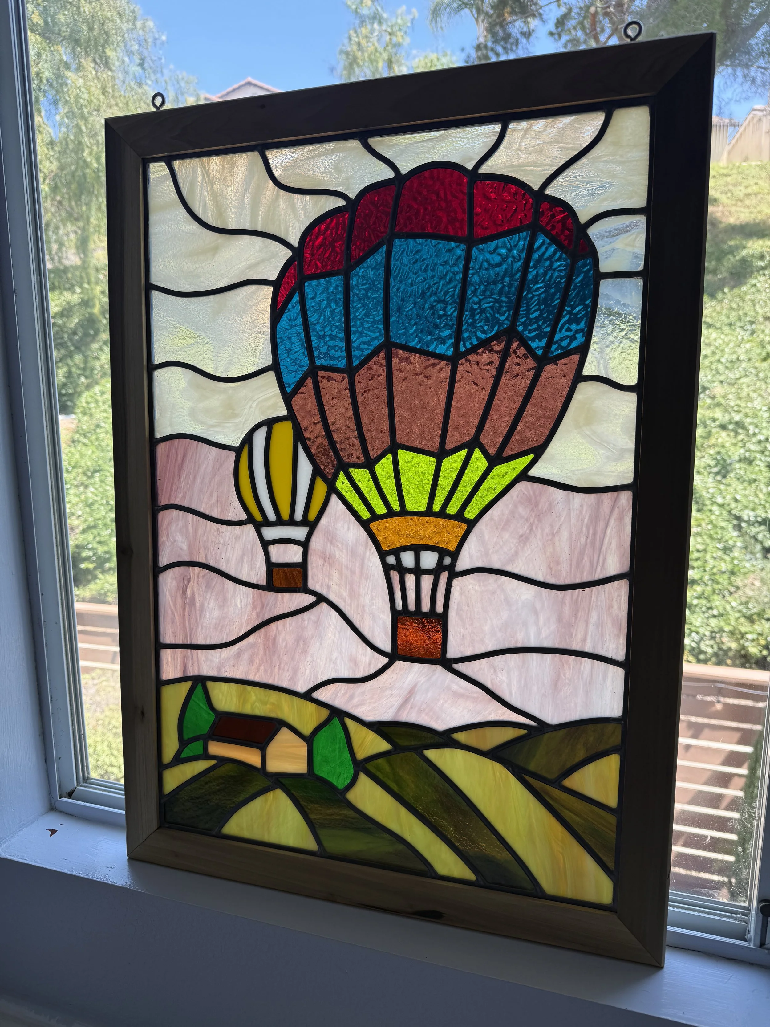 Colorful stained glass window depicting a hot air balloon with a blue and red balloon, yellow basket, and green landscape at the bottom, set in a wooden frame and displayed on a windowsill.