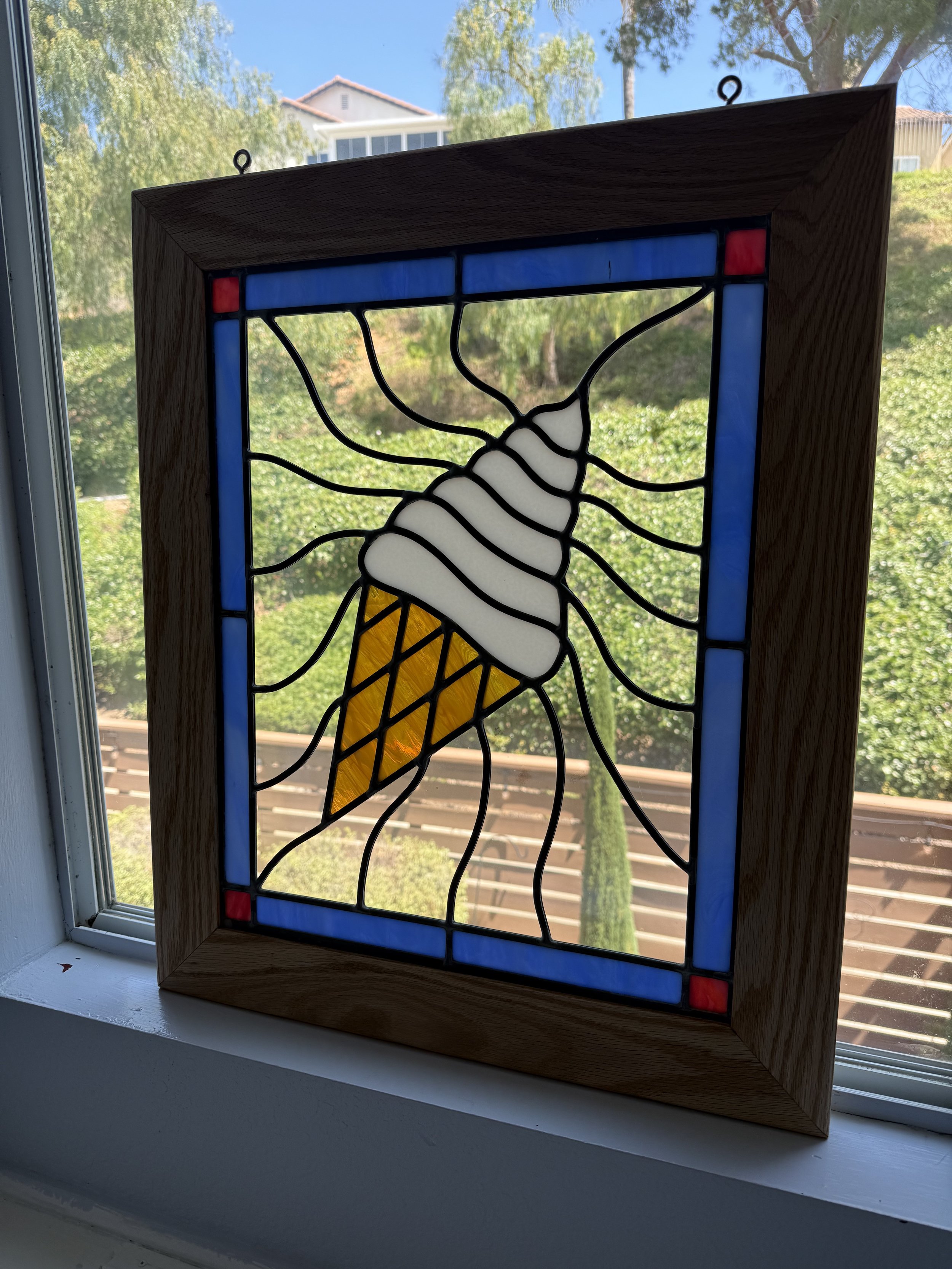 Stained glass window featuring an ice cream cone, framed with a wooden border, hanging in front of a window showing trees and houses outside.