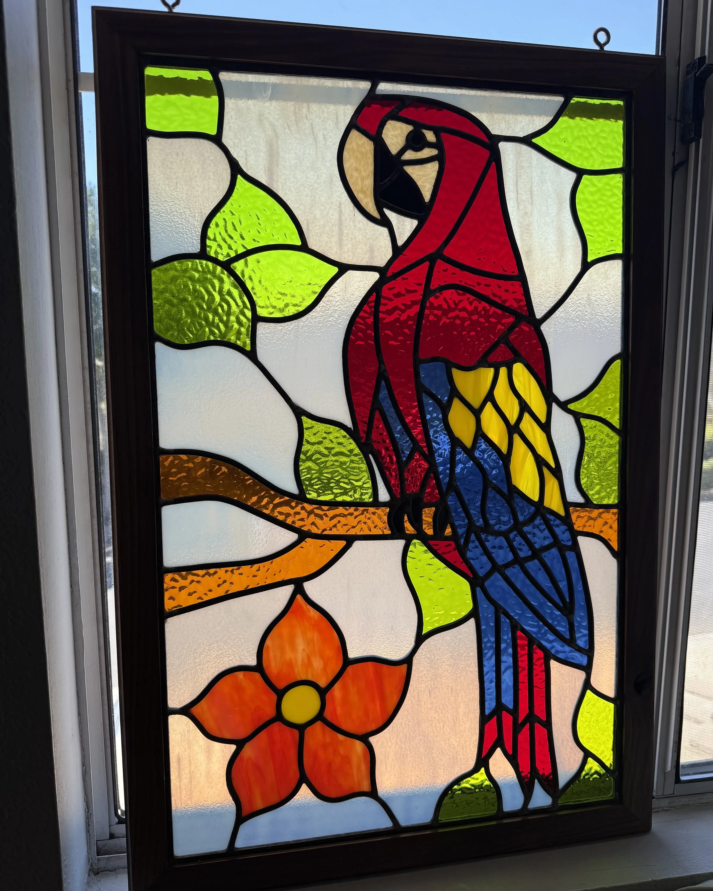 Colorful stained glass window featuring a toucan perched on a branch with leaves, flowers, and a sunrise or sunset in the background.