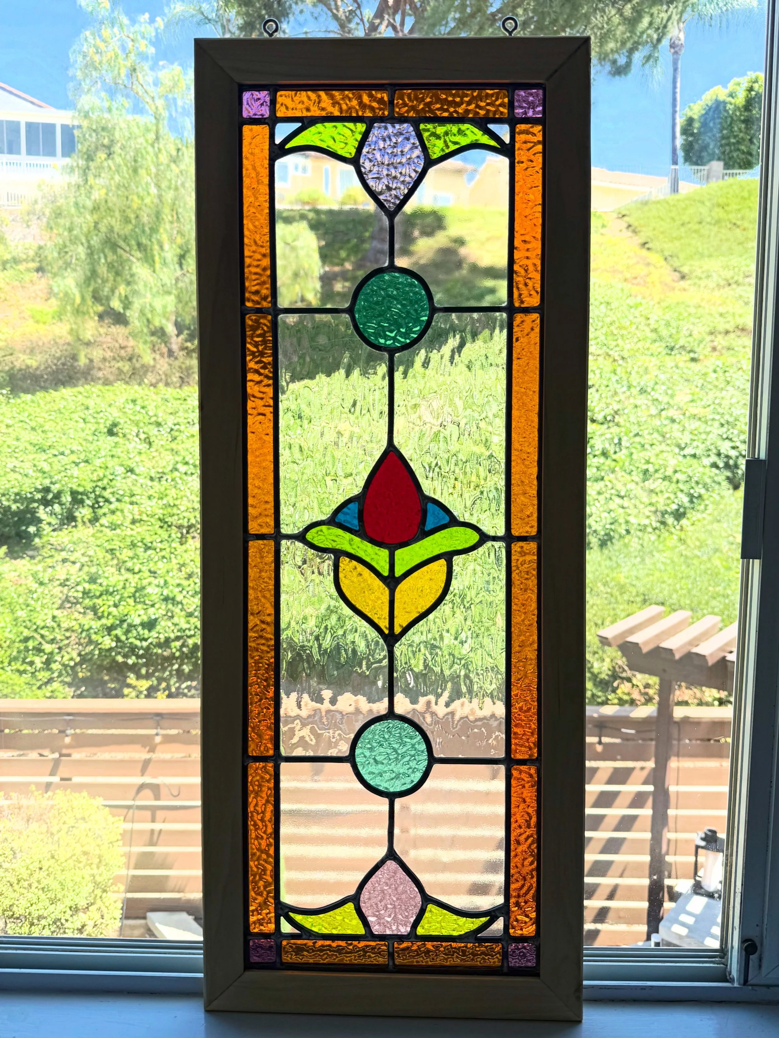 Colorful stained glass window with floral design and geometric patterns, framed and placed against a window showing an outdoor landscape.