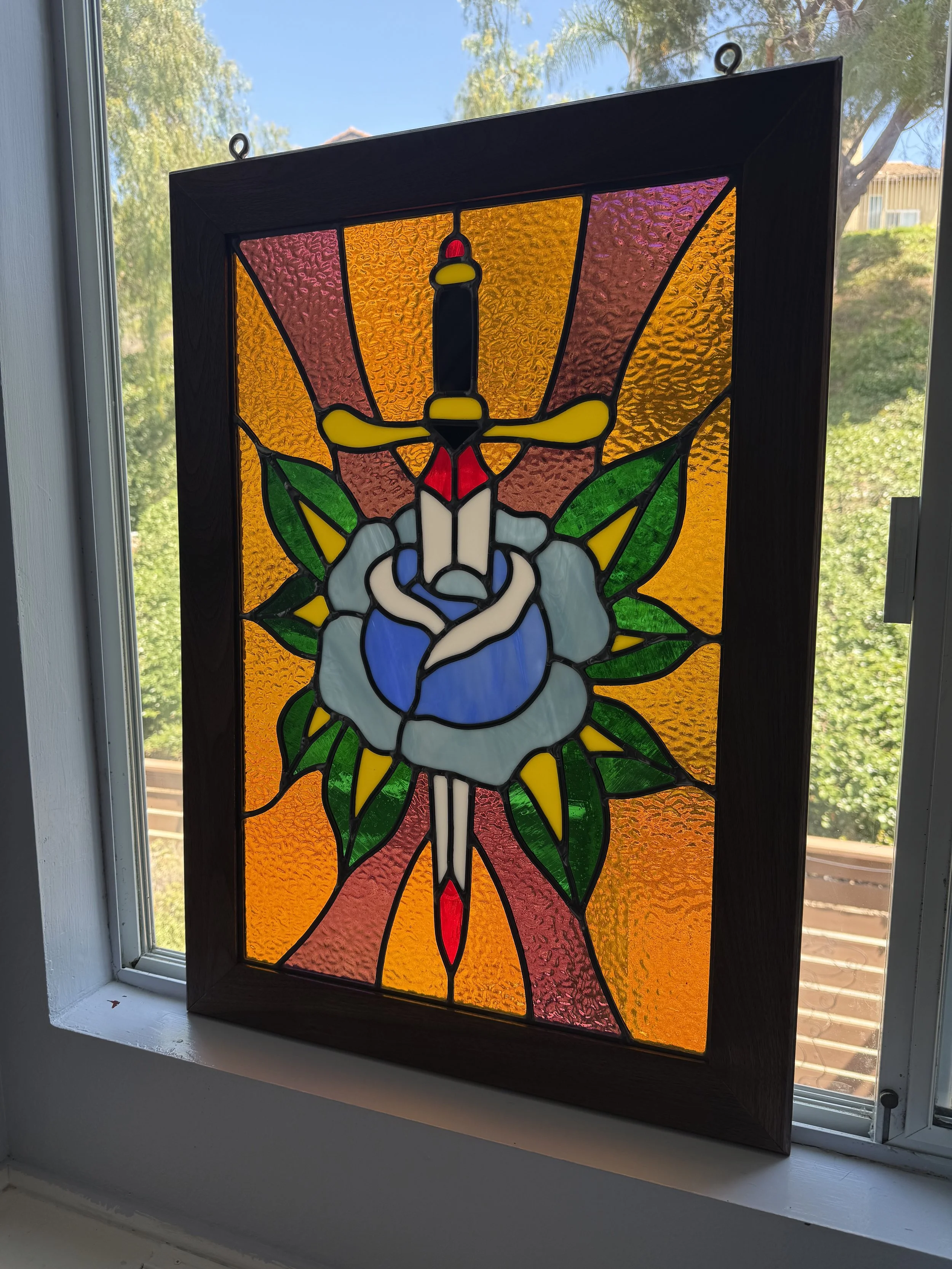 A stained glass window hanging on a wall shows a white and bluish rose, surrounded by colorful leaves with an orange and pink background, viewed in front of a window with trees and a blue sky outside.