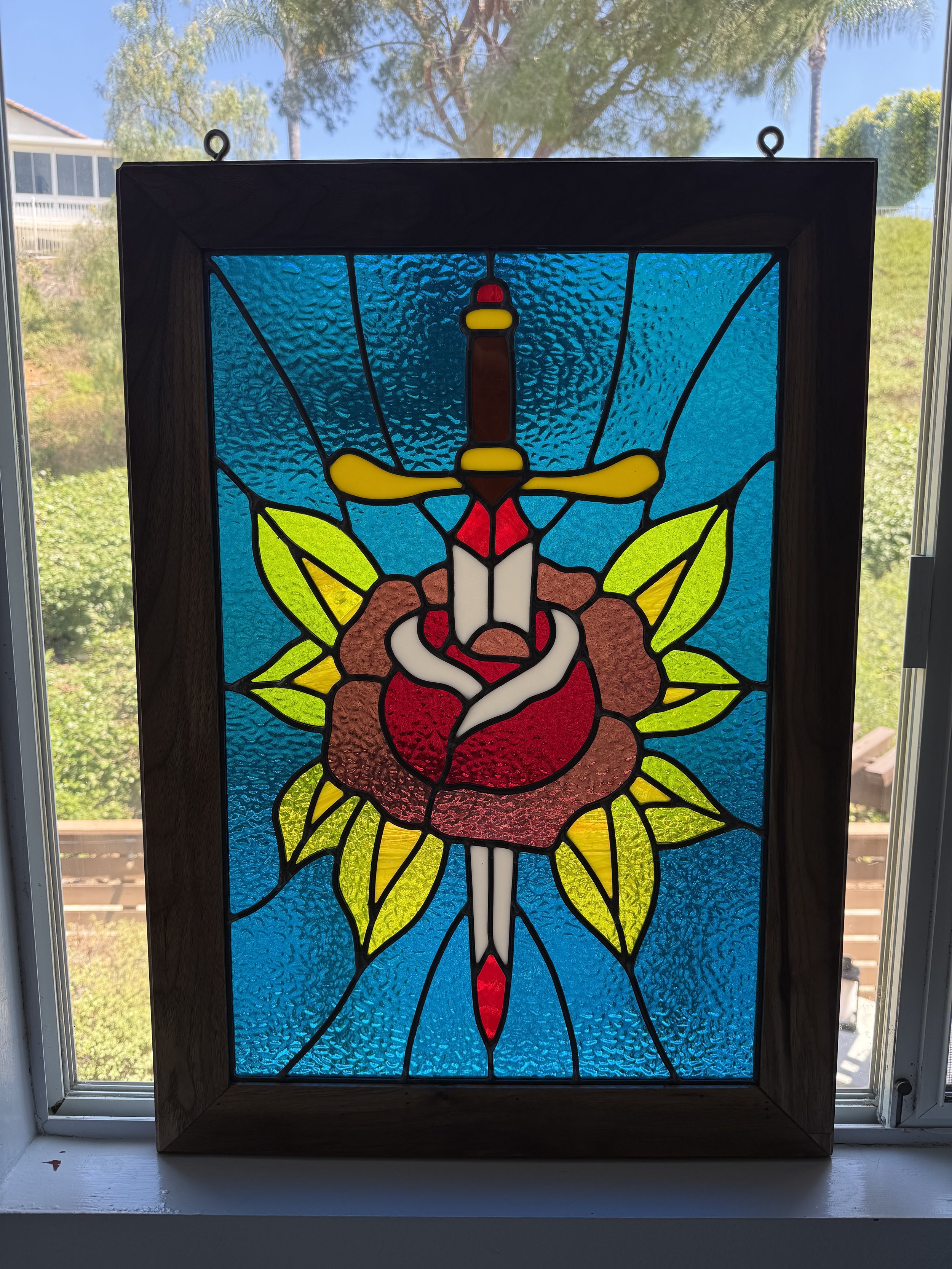 A stained glass window with a design of a vibrant red and white flower, green leaves, and a central vertical dagger, set against a blue textured background, framed by a wooden border, with a scenic outdoor view visible through the window.