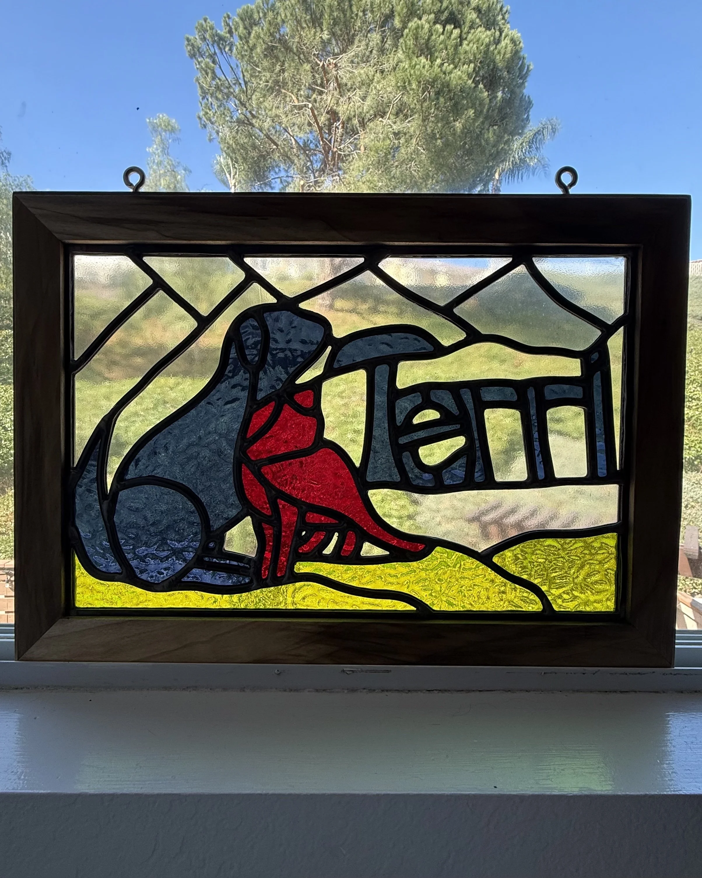 Colorful stained glass depicting a dog and cat with the name "TERRI", framed in wood, placed on a windowsill.