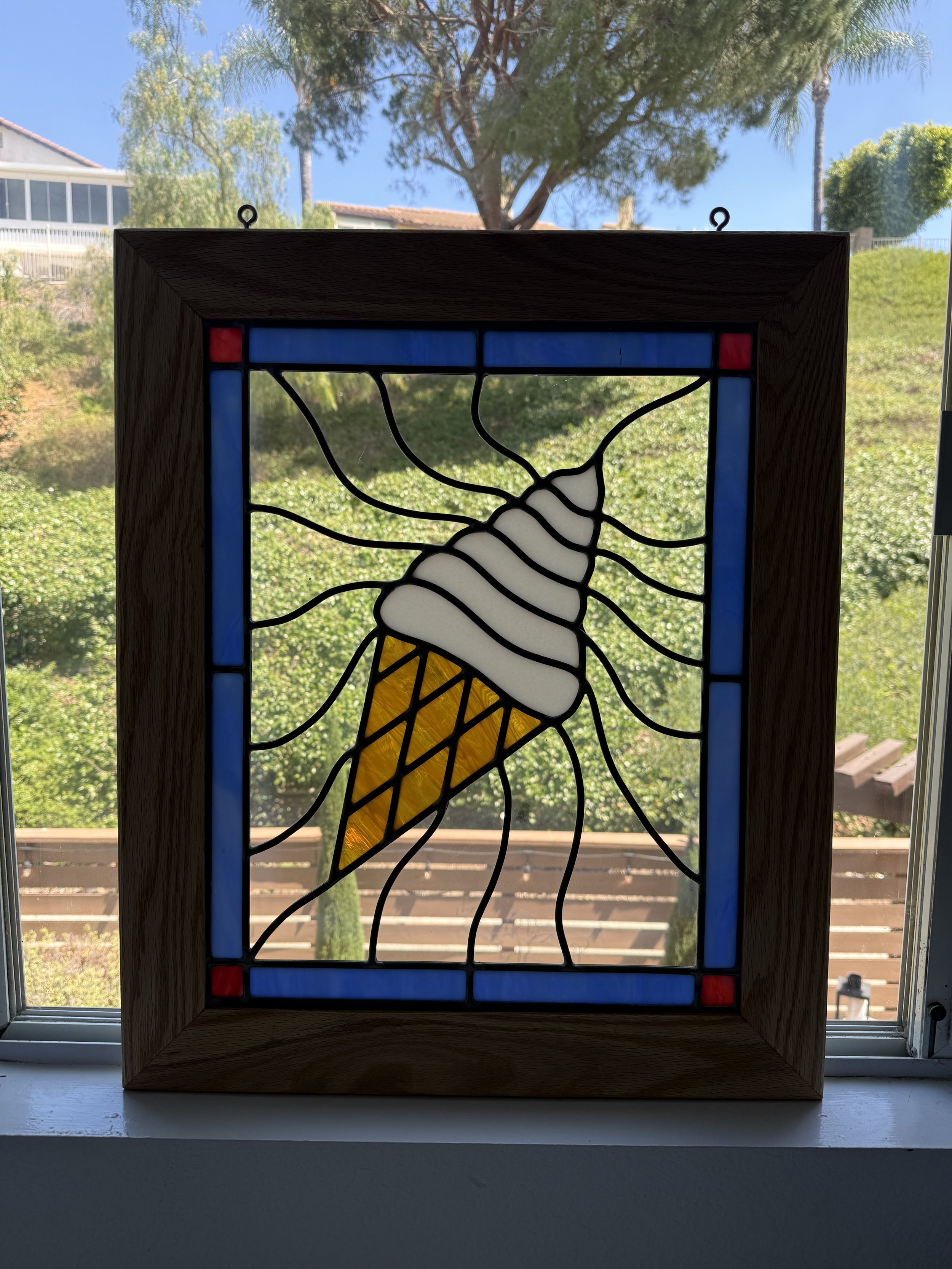 A stained glass artwork of an ice cream cone with black outlines, yellow and white body, located on a window with a view of trees and blue sky in the background.