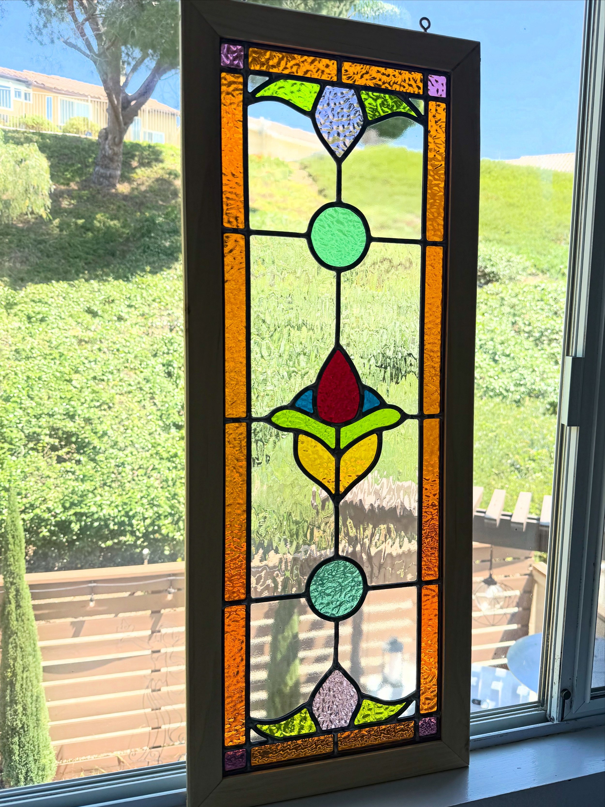 Colorful stained glass window with floral design, including green, purple, orange, red, yellow, and blue glass pieces, set in a white frame, seen against an outdoor garden scene with trees, bushes, and a wooden fence.