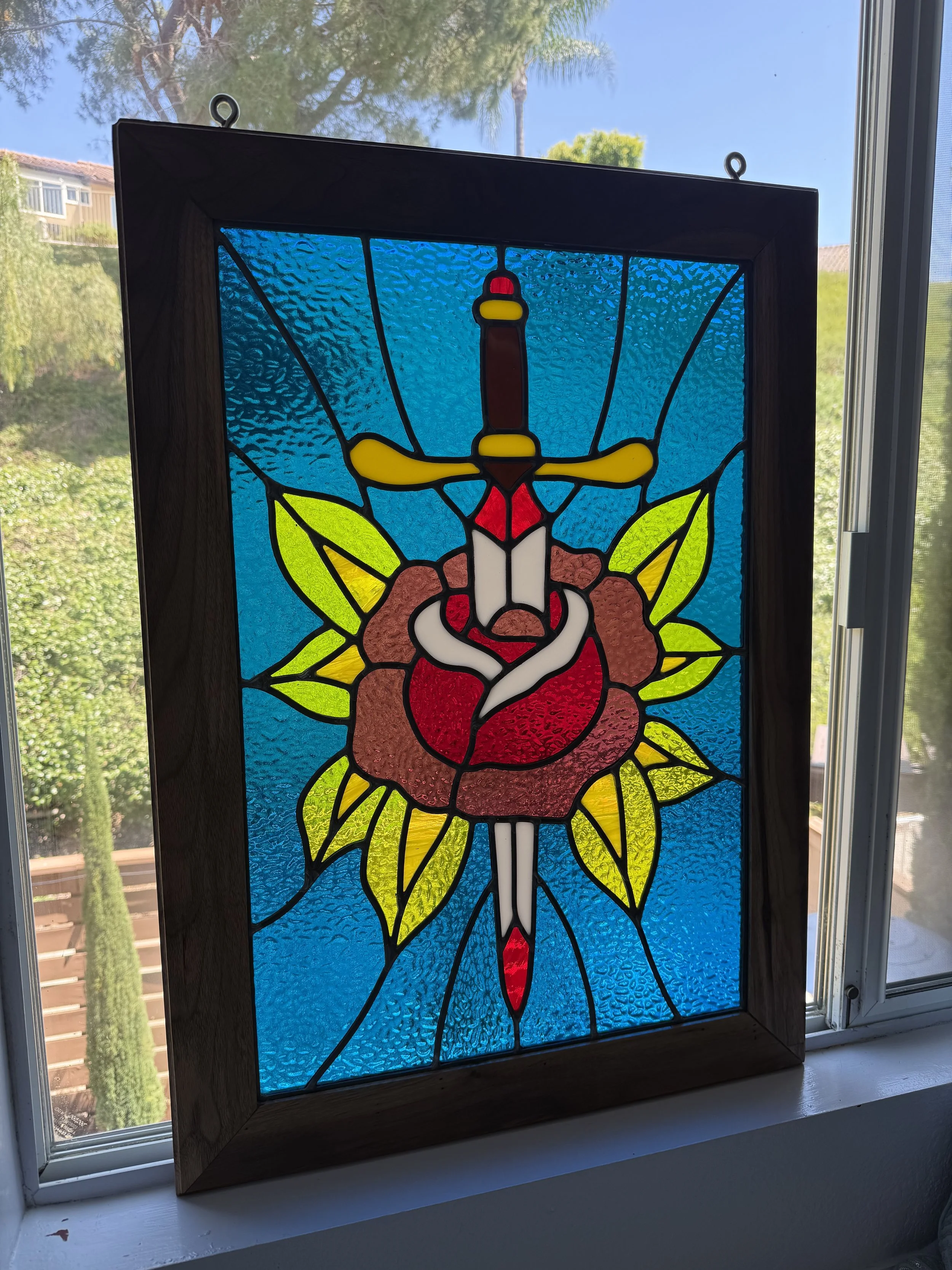 A stained glass window with a dagger and flower design, framed in wood, illuminated by sunlight coming through a nearby window.