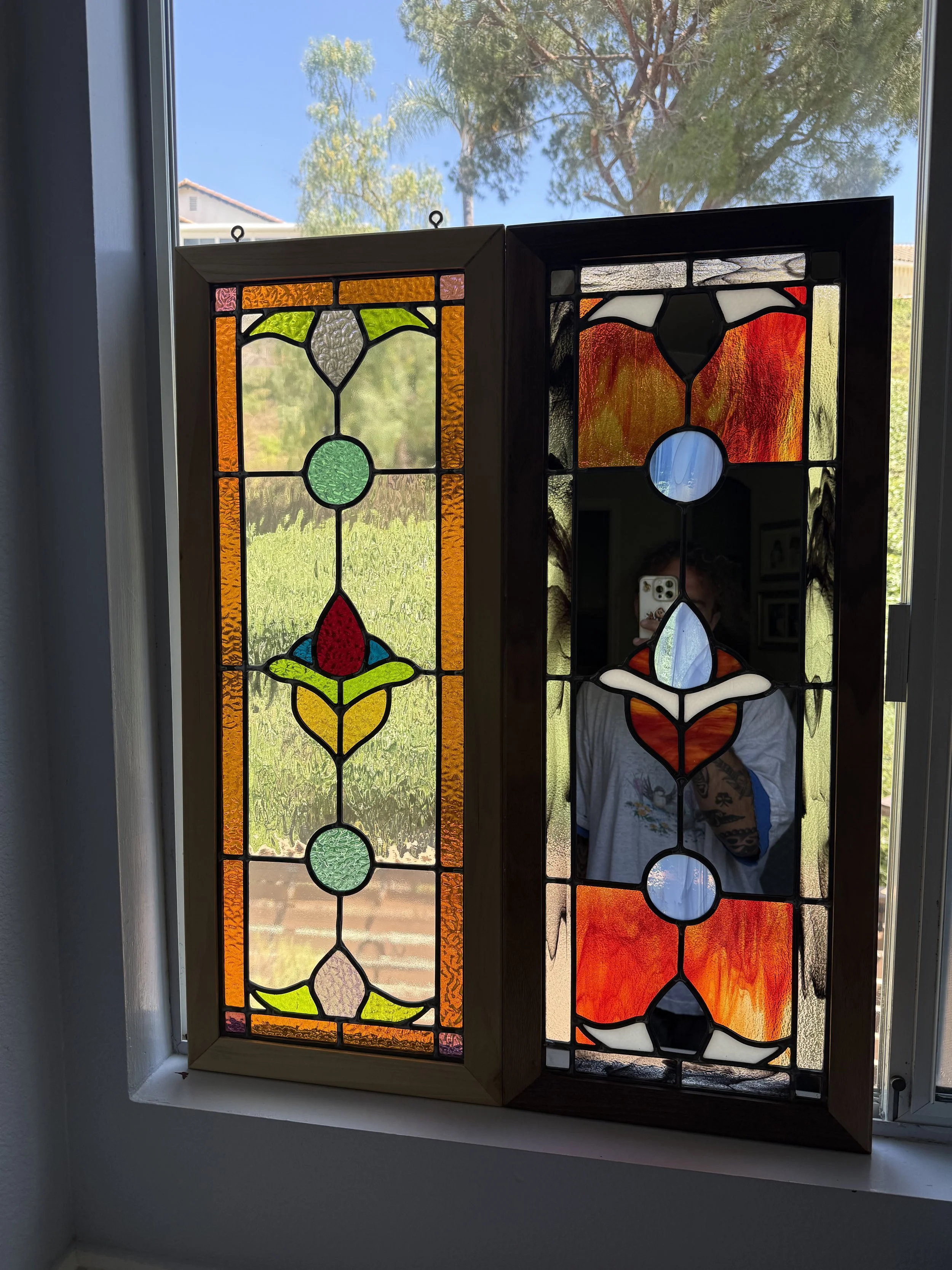 Colorful stained glass window panels with a floral and abstract pattern, framed in wood, installed in a window with trees and blue sky visible outside.