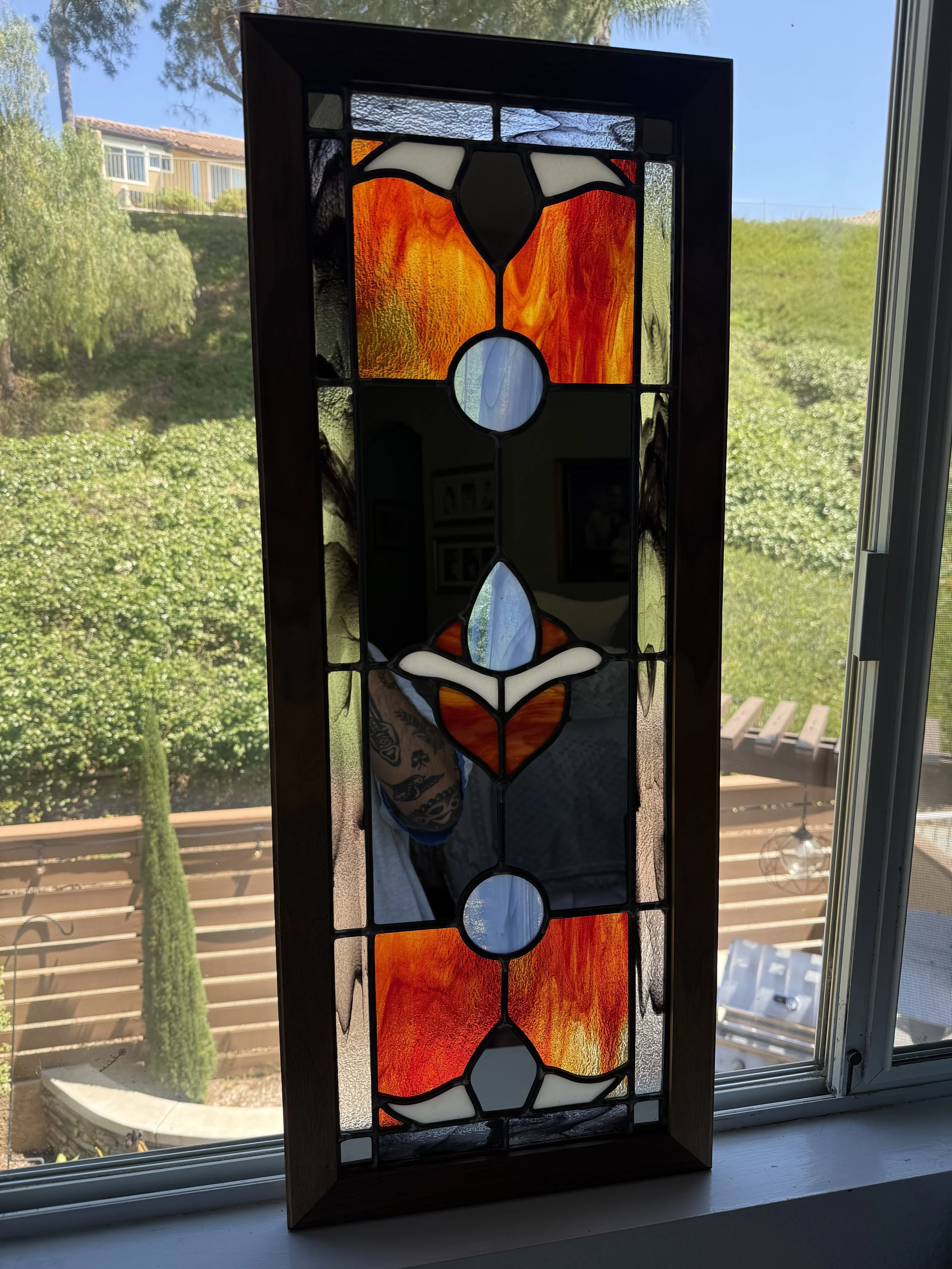 Stained glass window with abstract colored glass panels in white, orange, and red framed by a dark border, set in a wooden frame, backlit by sunlight.