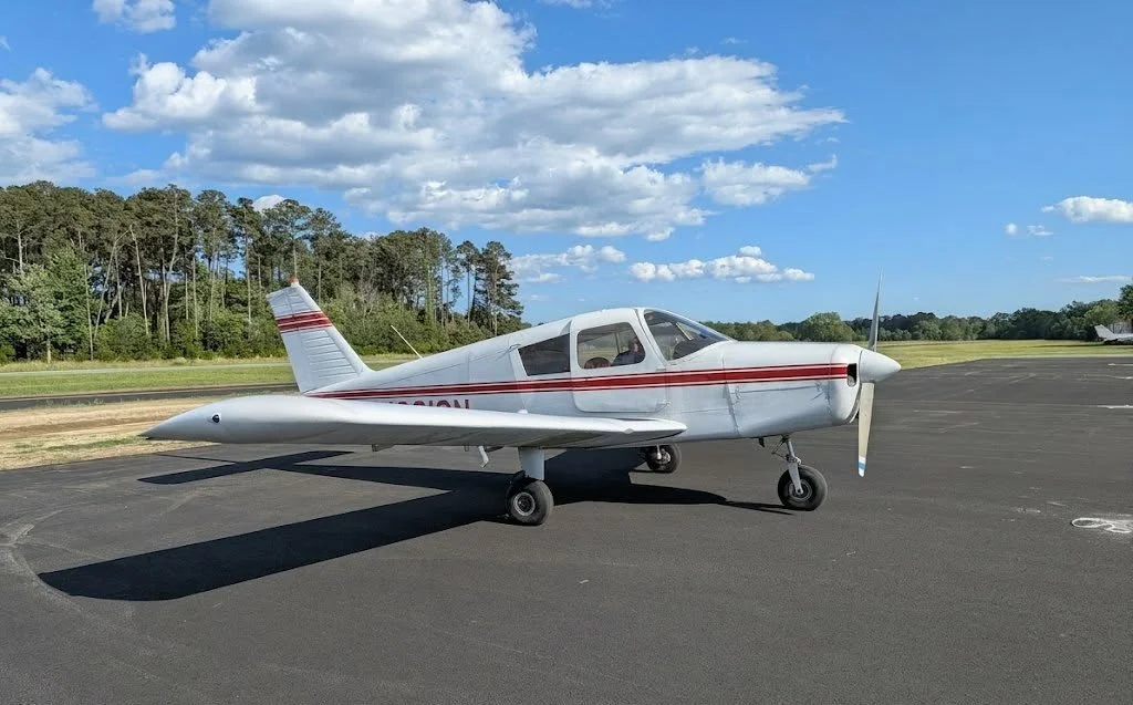Small white airplane with red stripes parked on runway under blue sky with clouds, surrounded by grassy fields and trees.
