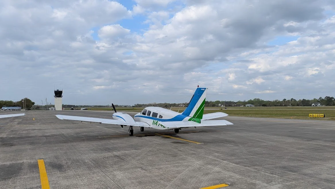 Small white propeller airplane with blue and green accents parked on an airstrip under partly cloudy sky.