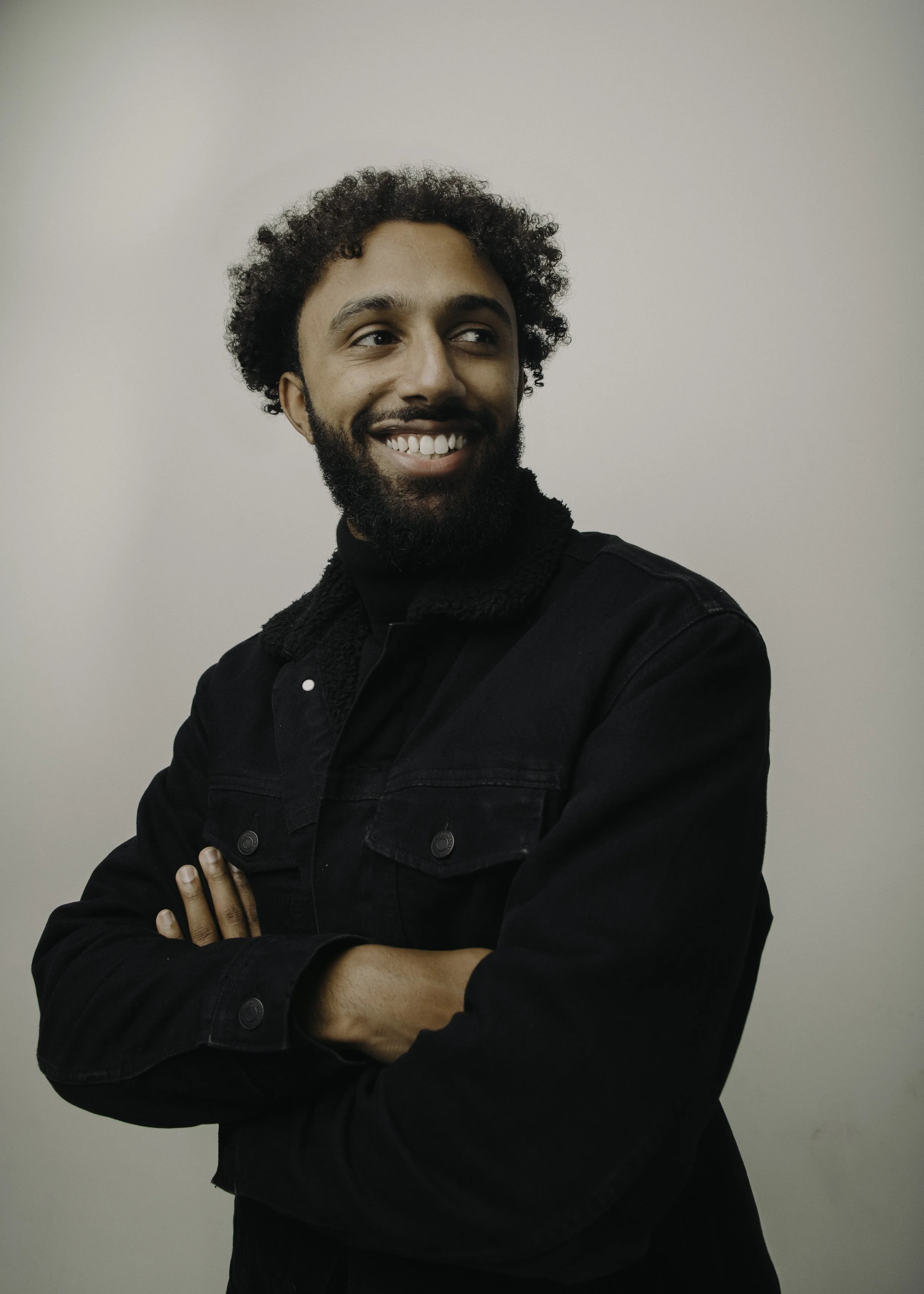 A smiling man with curly hair and a beard, wearing a black jacket with crossed arms, standing against a plain background.