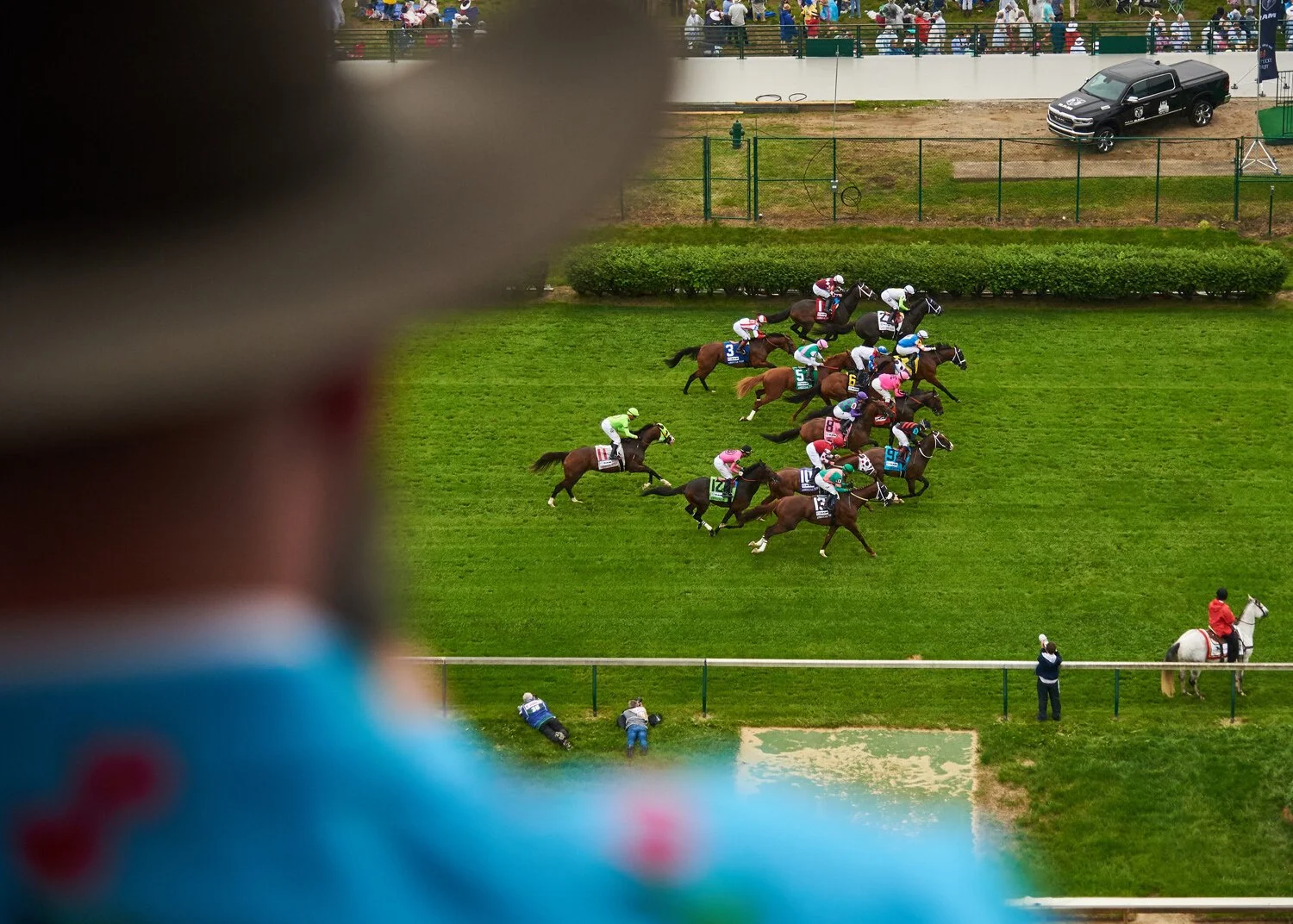 Horse race with jockeys racing on a green track, seen from above through a blurred figure in the foreground, with spectators and vehicles in the background.