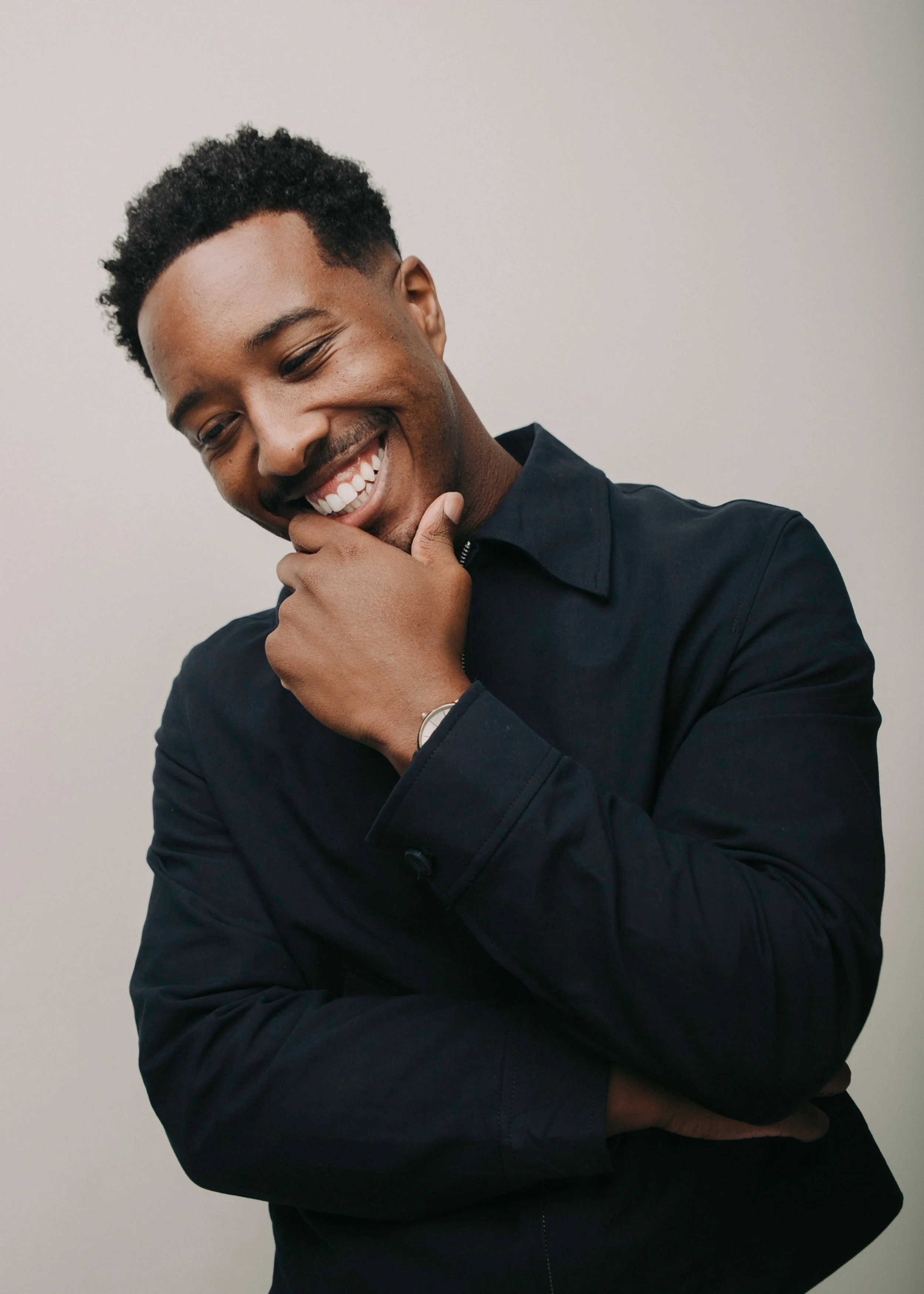 Young man smiling and looking down, wearing a black shirt, with hand on chin, against a plain background.