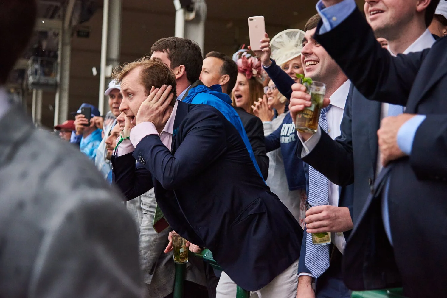 Group of people at a crowded outdoor event, many in formal attire, some holding drinks. The man in the foreground appears to be stressed or worried, while others around him are smiling and taking photos.
