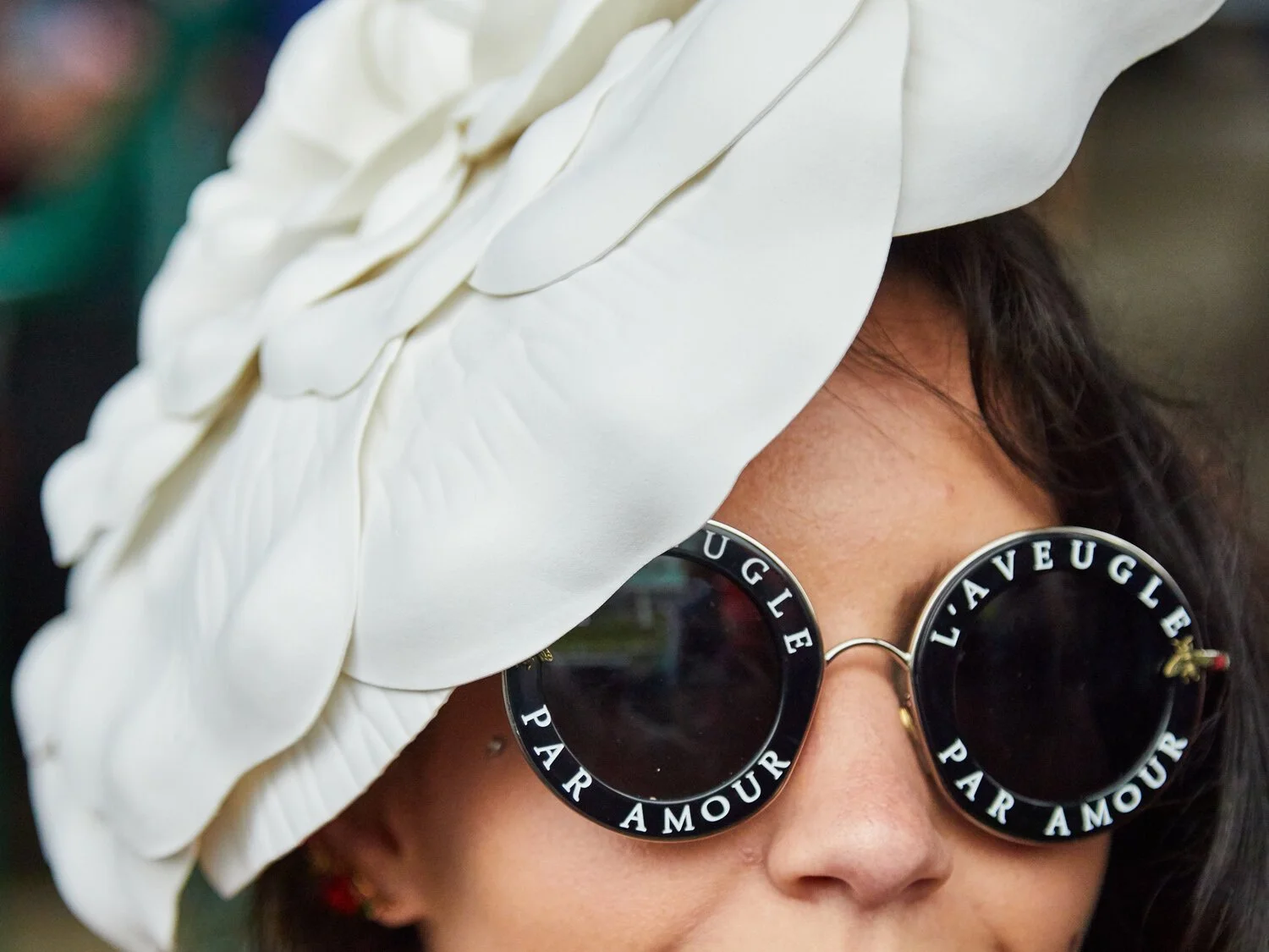 Close-up of a woman wearing large, round sunglasses with black frames that have the words 'L'AVUEUGLE' and 'PAR AMOUR' written on the rims. She is also wearing a wide, white hat decorated with large petals.