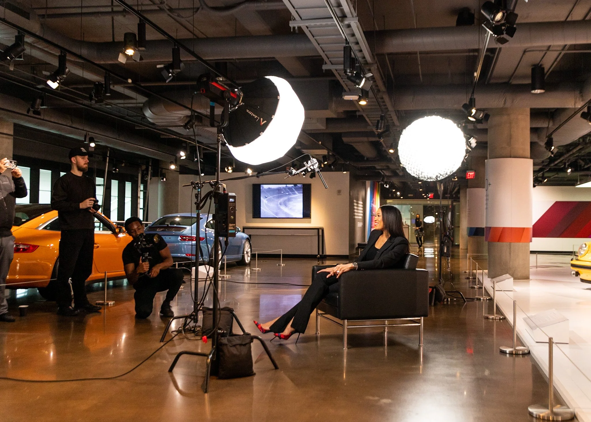 A woman in a black blazer and pants sitting on a black chair in a car museum, being filmed by a camera crew with lighting equipment. Several cars are displayed behind her with showcases and a large monitor on the wall.