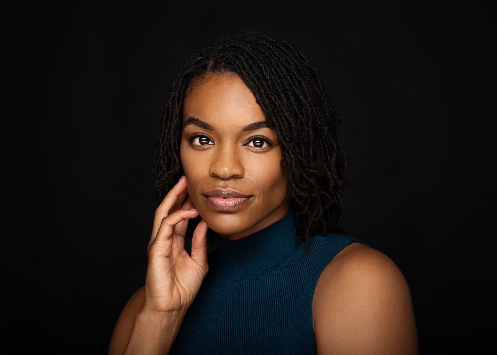 Portrait of a woman with shoulder-length black curly hair, wearing a sleeveless navy blue top, posing against a black background.