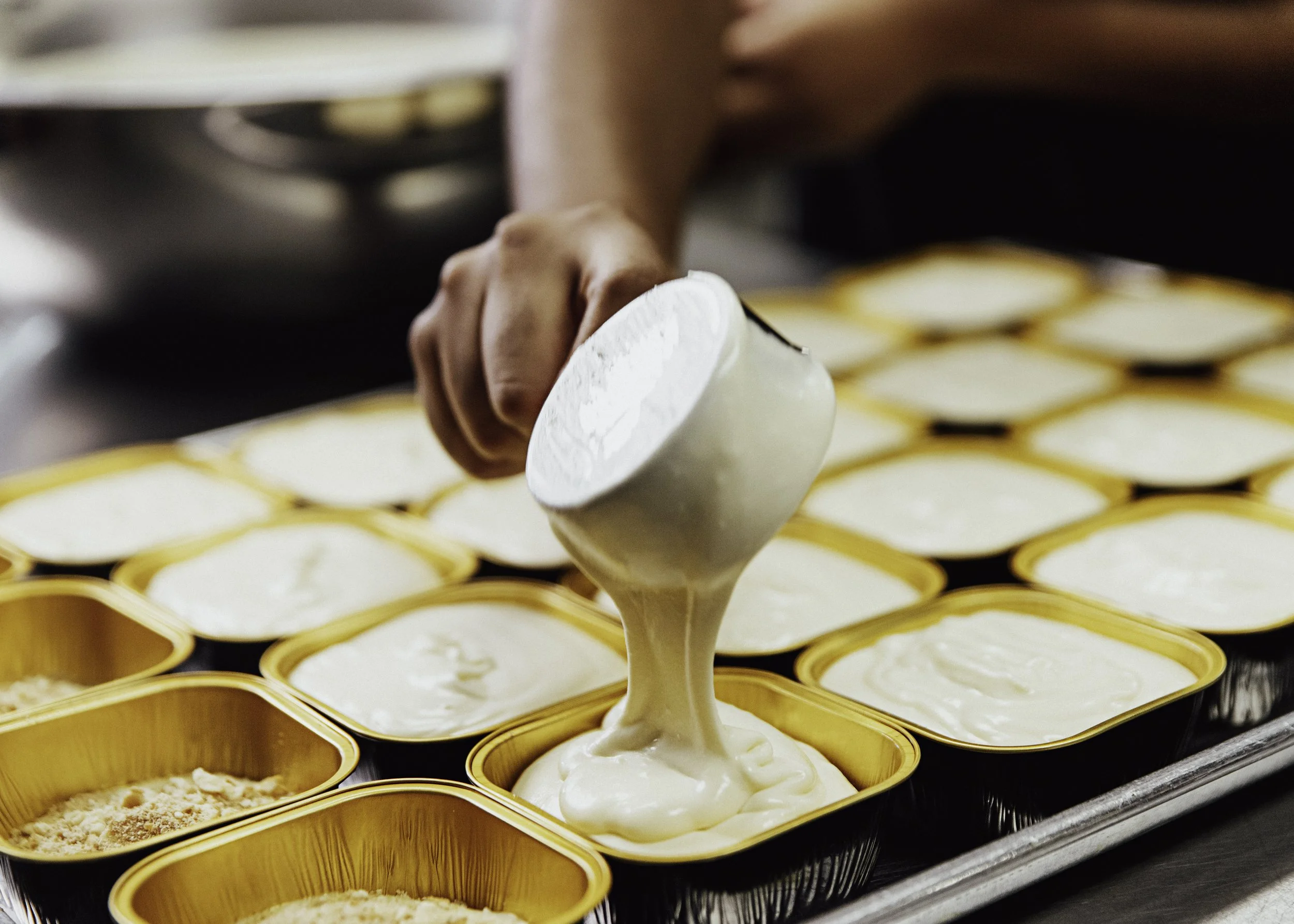 Person pouring creamy white dessert into small square yellow aluminum containers filled with similar desserts, with some containers topped with a crumb mixture, on a silver tray.