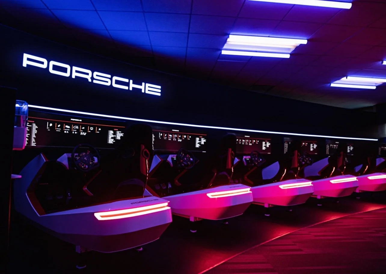 Indoor racing simulators with Porsche branding, illuminated with pink and purple neon lights, arranged in a row, with digital screens and racing seats.