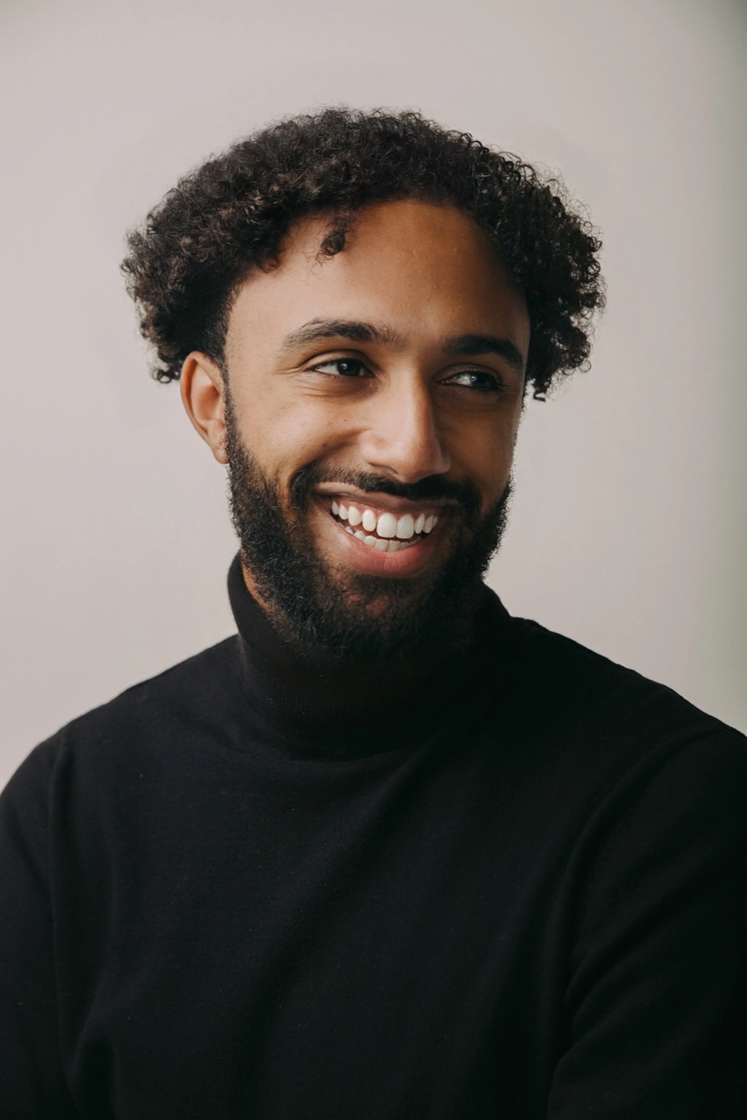 A smiling man with curly hair and a beard wearing a black turtleneck, looking to his right against a plain light background.