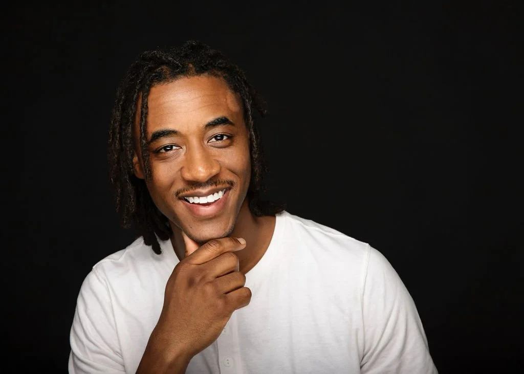 A young man with dreadlocks smiling and resting his chin on his hand against a black background.