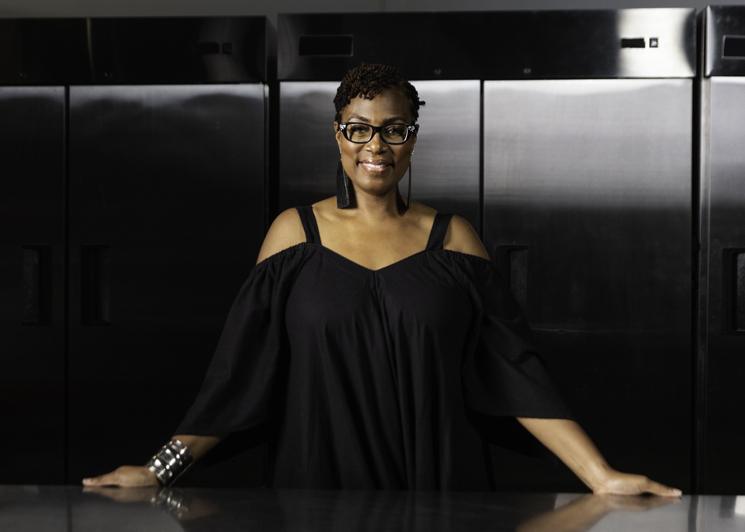 A woman standing in front of stainless steel kitchen appliances, smiling at the camera, wearing glasses, a black off-shoulder dress, and a silver cuff bracelet.