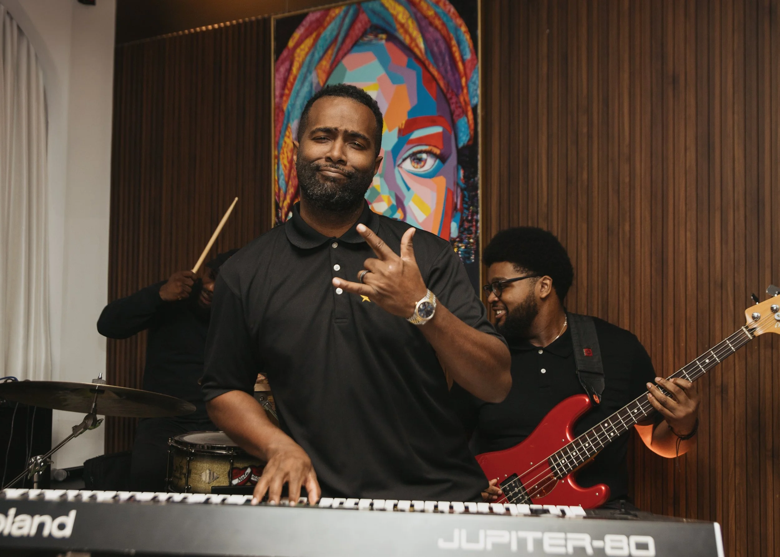 Musicians performing indoors with a colorful portrait of a woman in the background. The man in the foreground is playing keyboard, making a gesture with his hand and wearing a black polo shirt. Another man behind him is playing a red bass guitar, and