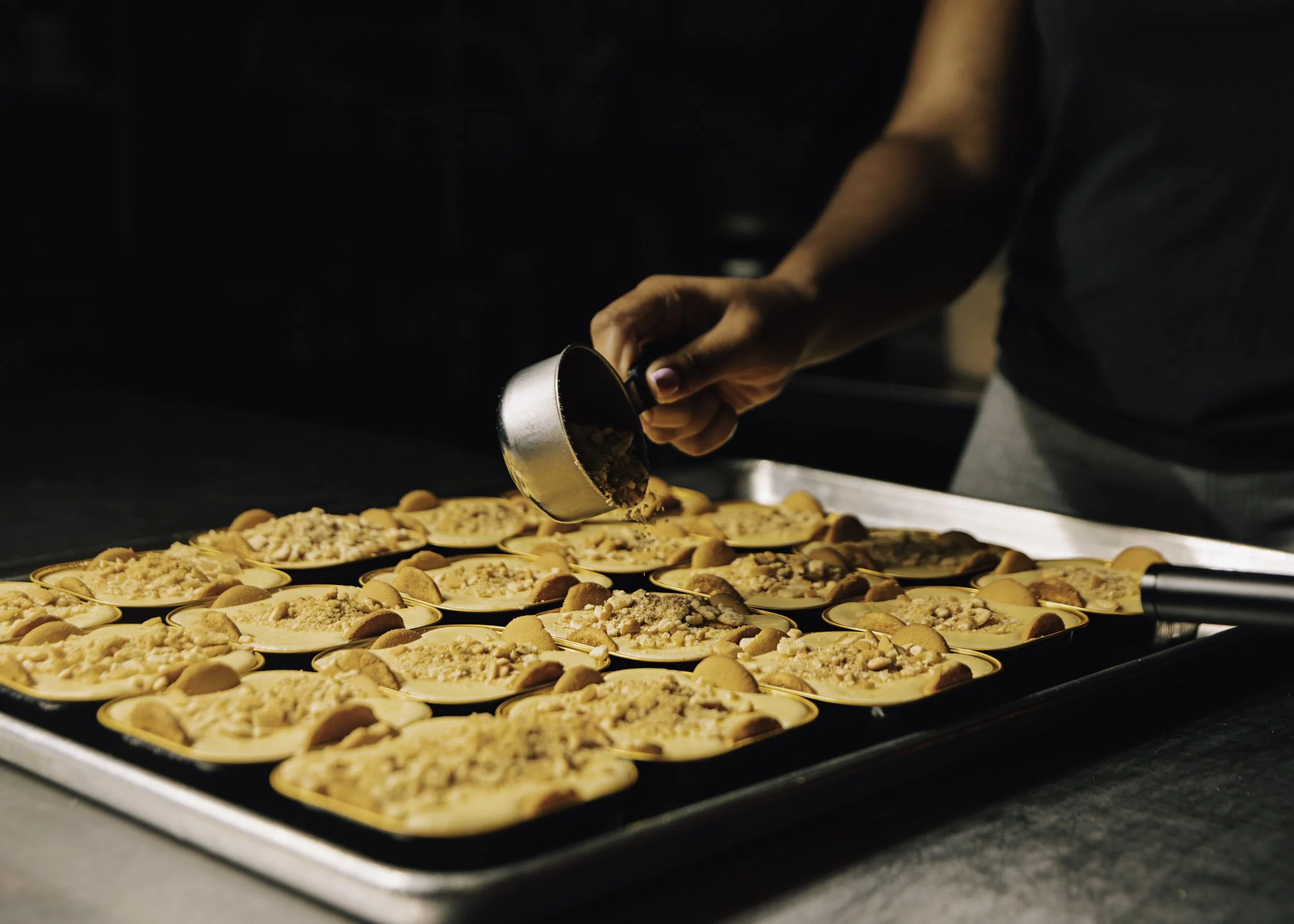 A person sprinkling crushed nuts or crumbs onto mini dessert cups on a baking tray.