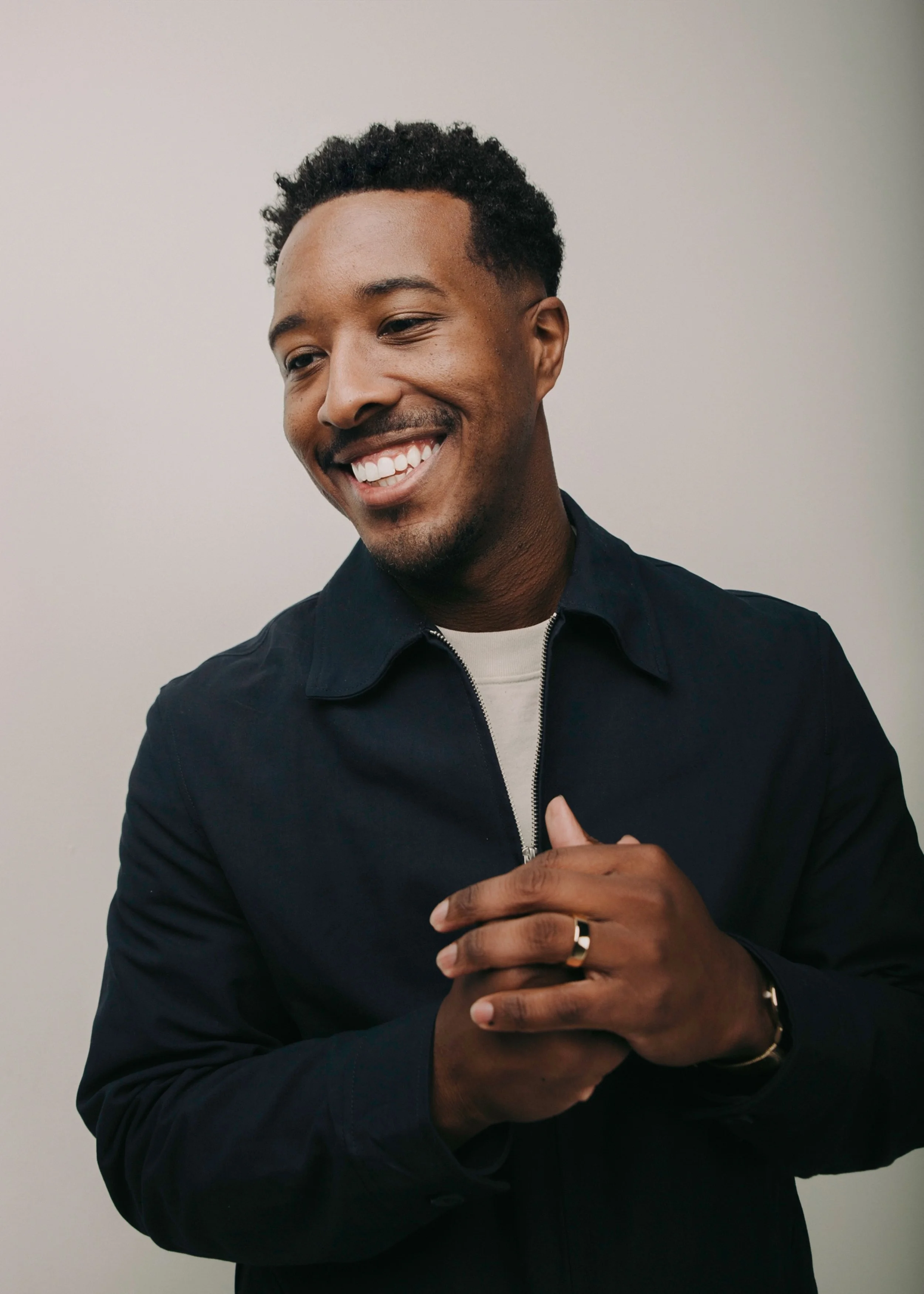 A smiling man with short curly hair, wearing a dark jacket and a white shirt, standing against a plain background.