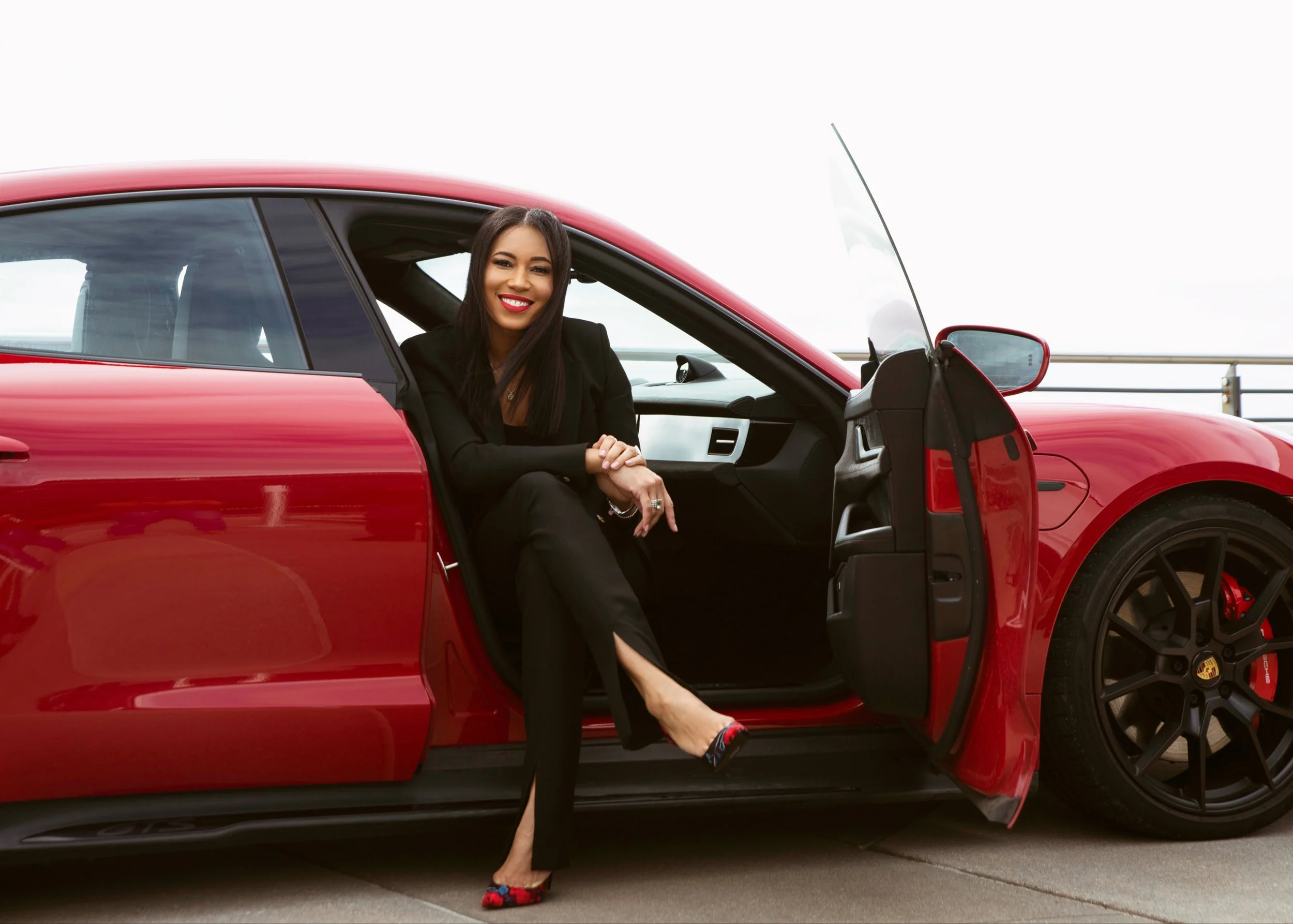 A woman with black hair and red lipstick sitting in a red sports car, smiling at the camera. She is wearing a black suit and high heels, with her legs crossed outside the car door.
