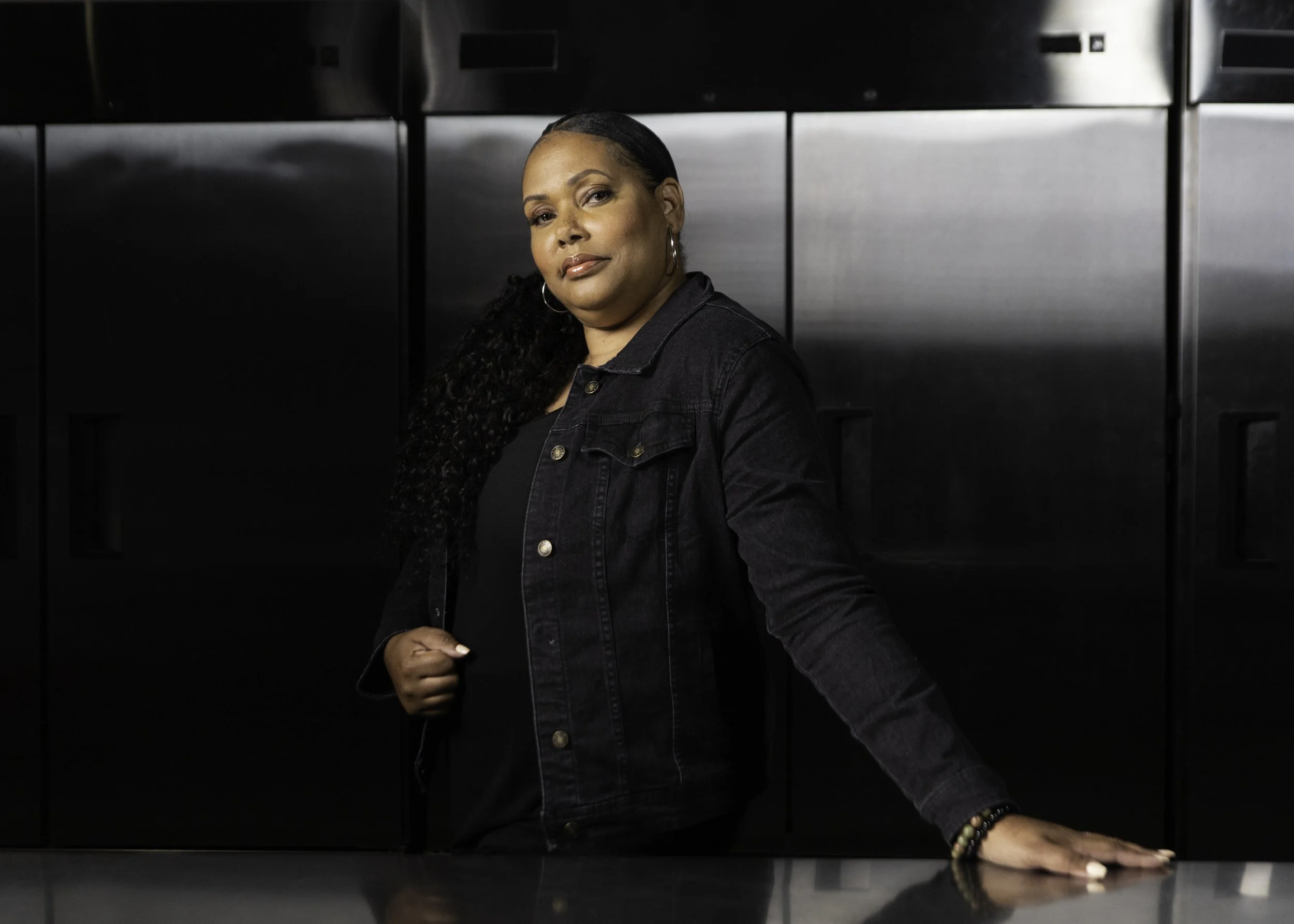 A woman with dark, curly hair wearing a black shirt and jeans, standing in front of black appliances or lockers, looking confidently at the camera.