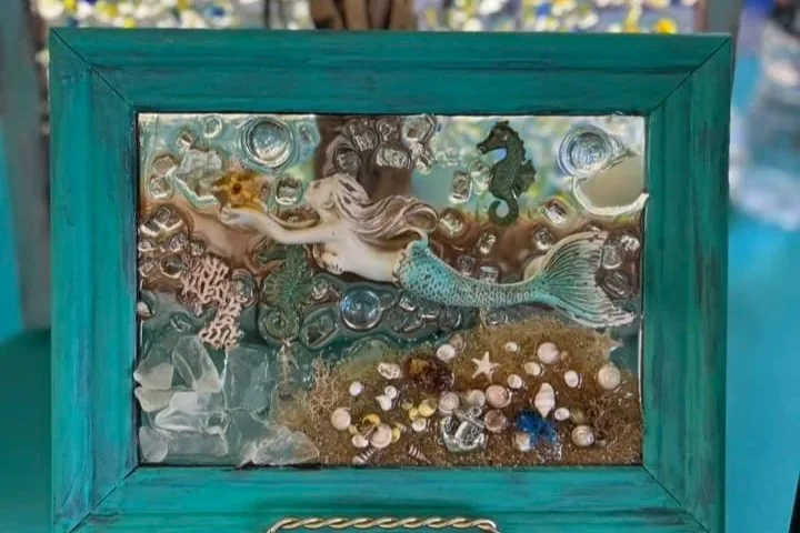 A framed underwater scene with a mermaid figurine, surrounded by seashells, rocks, and water-themed decorations.