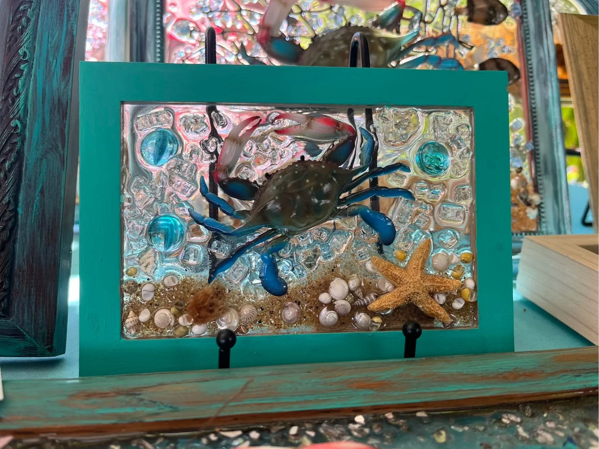 A glass display box with a blue frame contains a decorative arrangement including a plastic crab, a starfish, small shells, and sand, all enclosed in a clear glass panel.