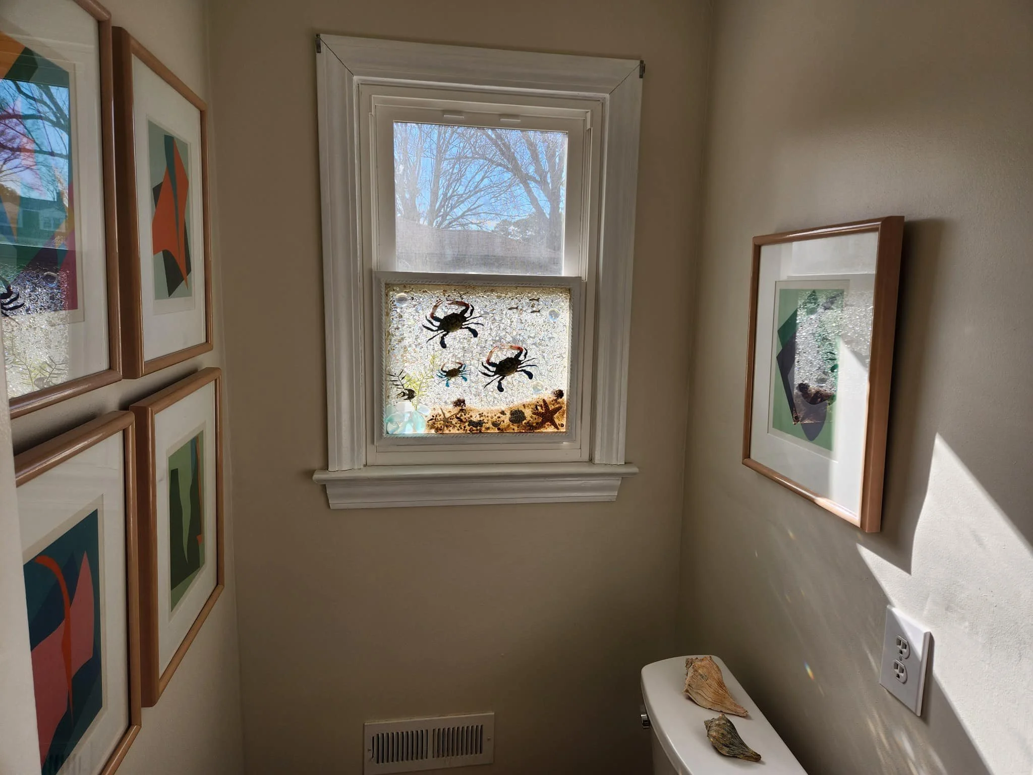 A small room or corner with framed abstract art on the walls, a window with decorative stickers of crabs, a starfish, and seaweed, and shells placed on a white surface near an electrical outlet