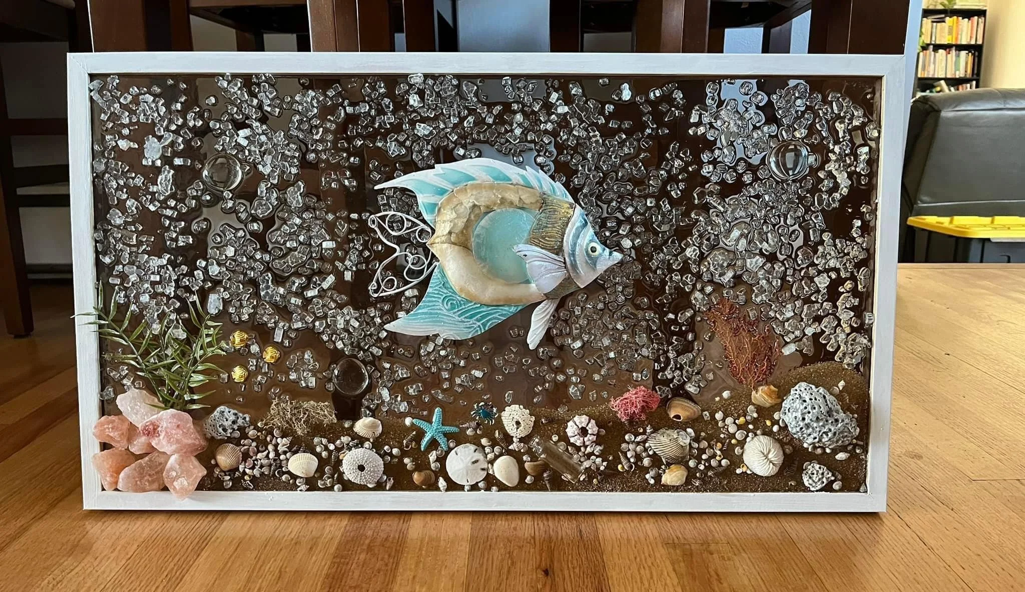 A decorative framed underwater scene with a large fish sculpture, seashells, coral, starfish, rocks, and small plants on a sandy surface.