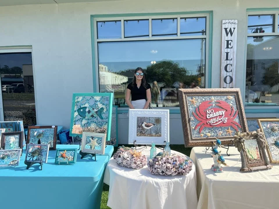 Outdoor art and craft booth with framed seashell and coastal artwork, decorative shells, and marine-themed figurines, with a smiling woman standing behind the display.