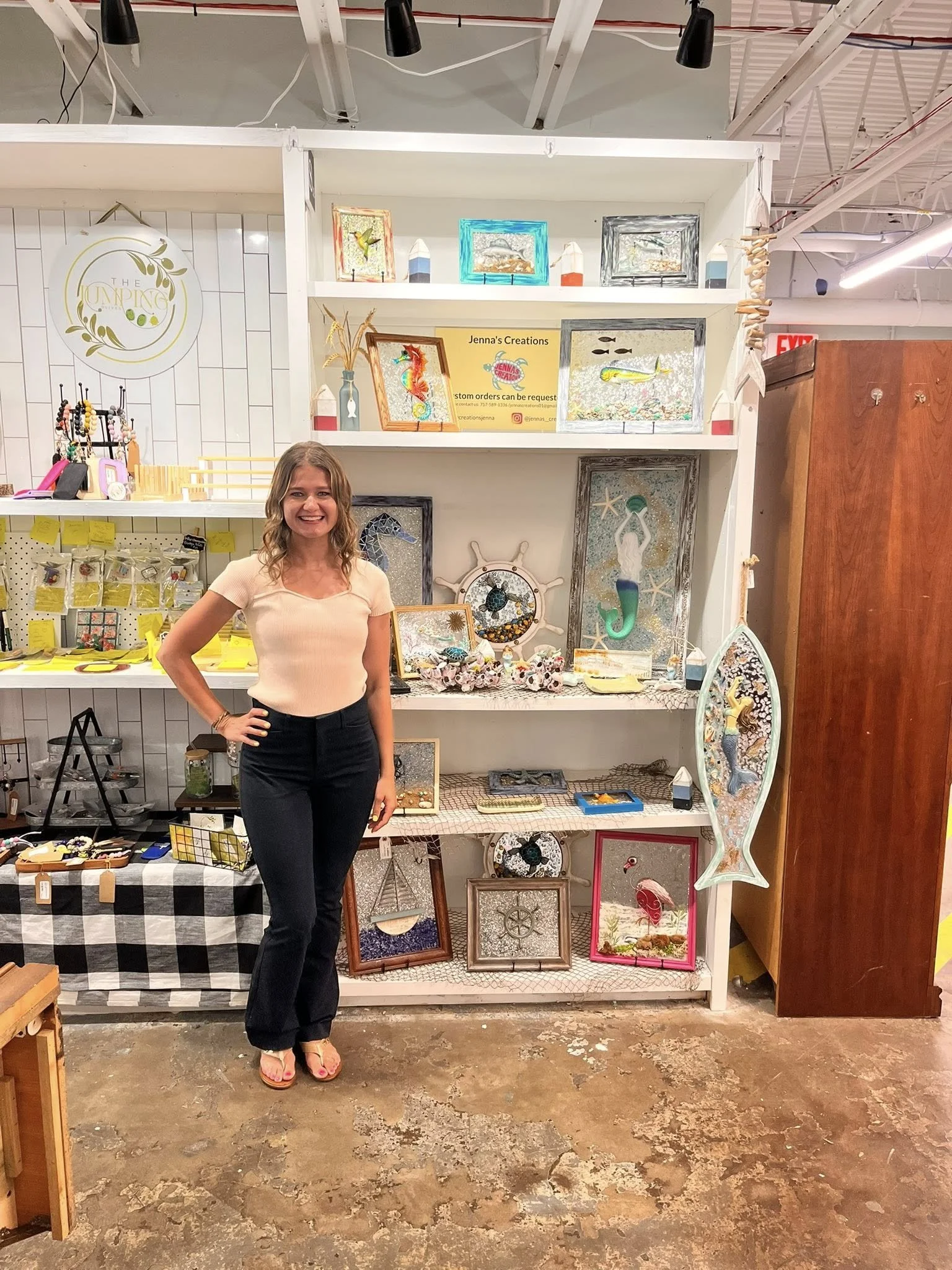 A woman standing in front of a white shelving unit filled with handcrafted art and decor, smiling at the camera.