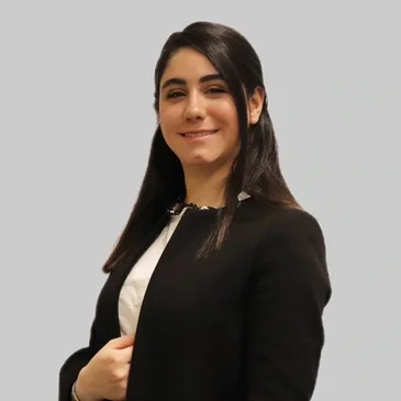 A young woman with long dark hair smiling, wearing a black blazer and white shirt, standing against a plain light gray background.