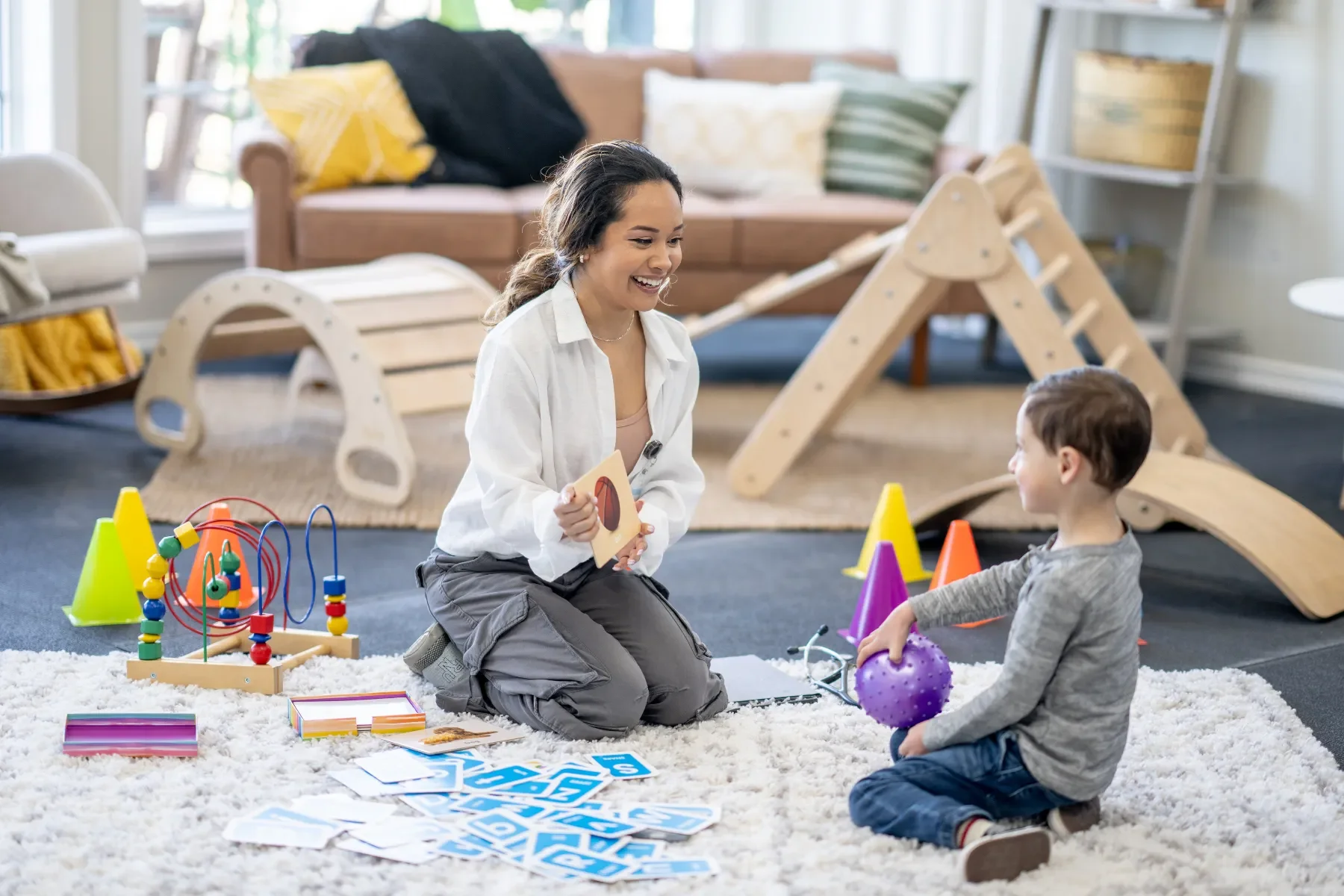 A woman playing a game with a young boy on a white carpet in a playroom. The woman is holding a card and smiling at the boy, who is holding a purple textured ball. The playroom has wooden playground equipment, colorful cones, and various toys and games scattered around.