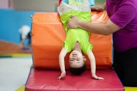 Child performing a backbend on gym mats, assisted by an adult, in a gym setting.