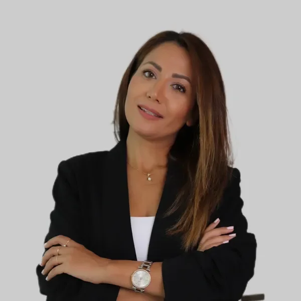 A woman with long brown hair, wearing a black blazer, white top, and a gold watch, standing against a plain light gray background.