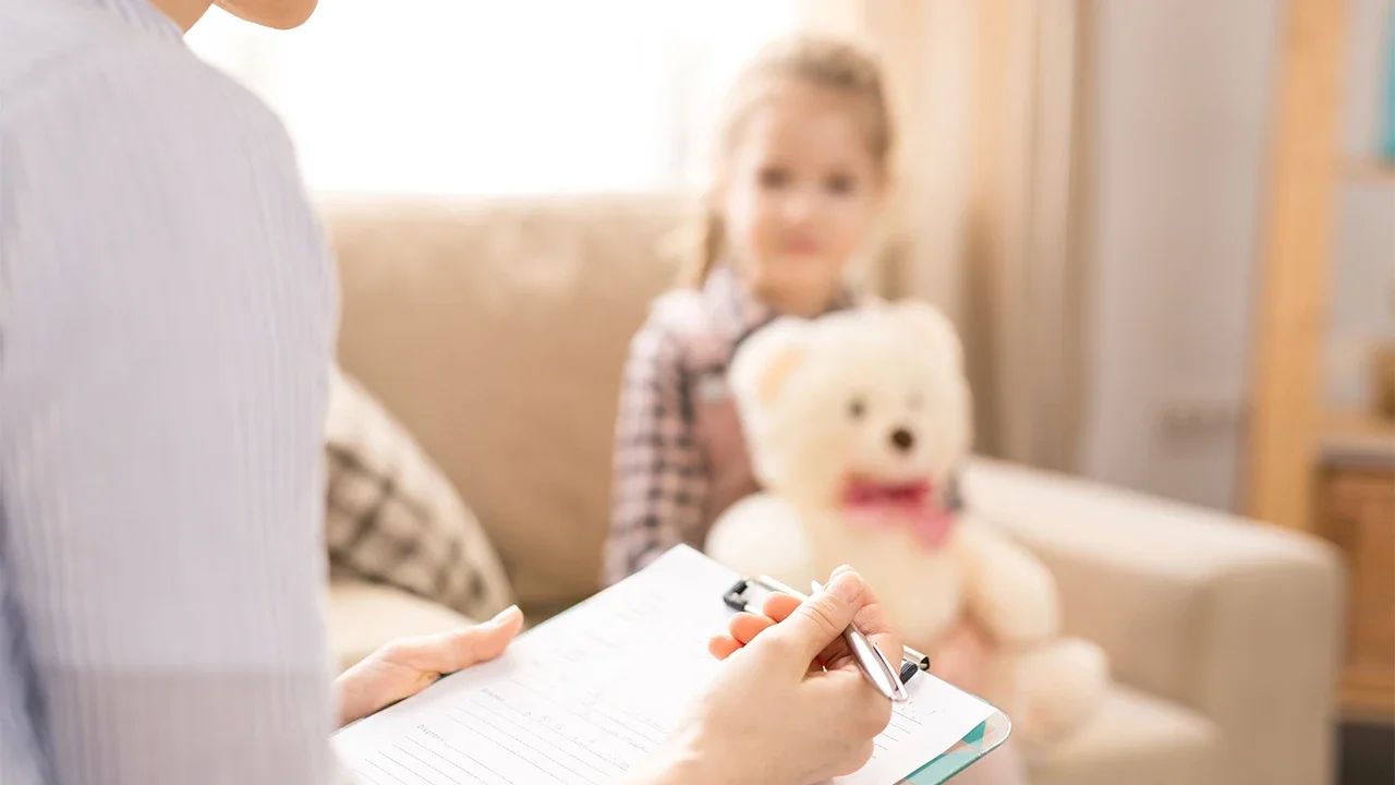 Child sitting on a couch holding a teddy bear, with an adult writing on a clipboard in the foreground