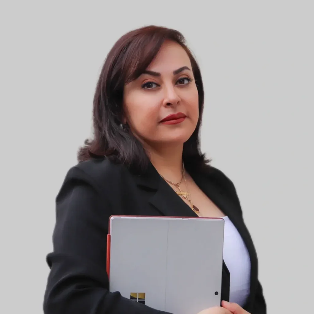A woman with shoulder-length dark hair, wearing a black blazer and white top, holding a closed laptop in front of her chest, standing against a plain light-colored background.