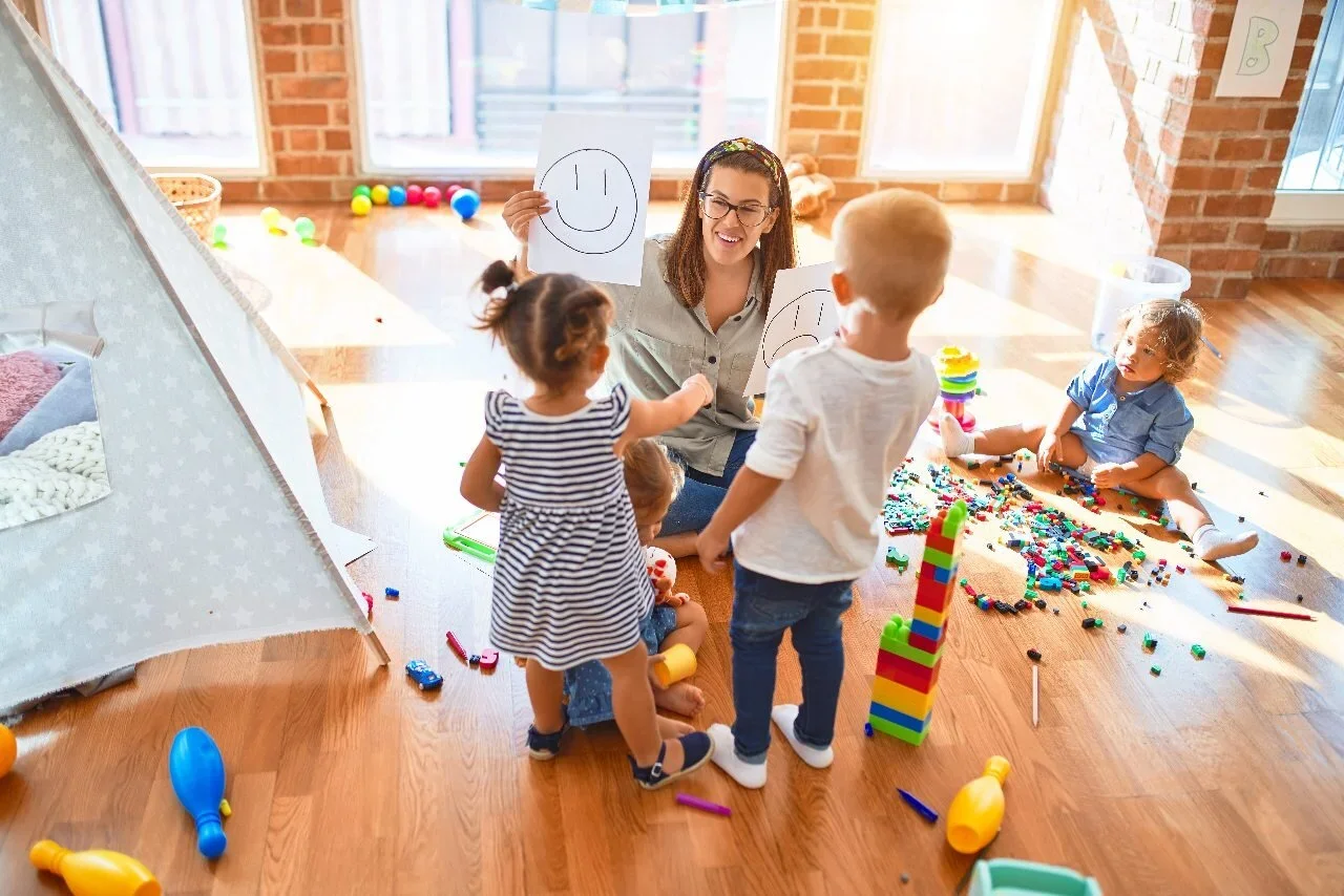A woman teaches children to draw smiley faces in a bright playroom with toys and scattered building blocks.