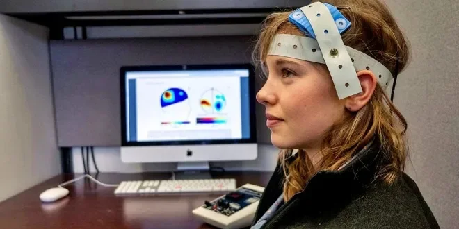 Woman wearing an EEG cap in a lab with a computer displaying brain wave graphs.